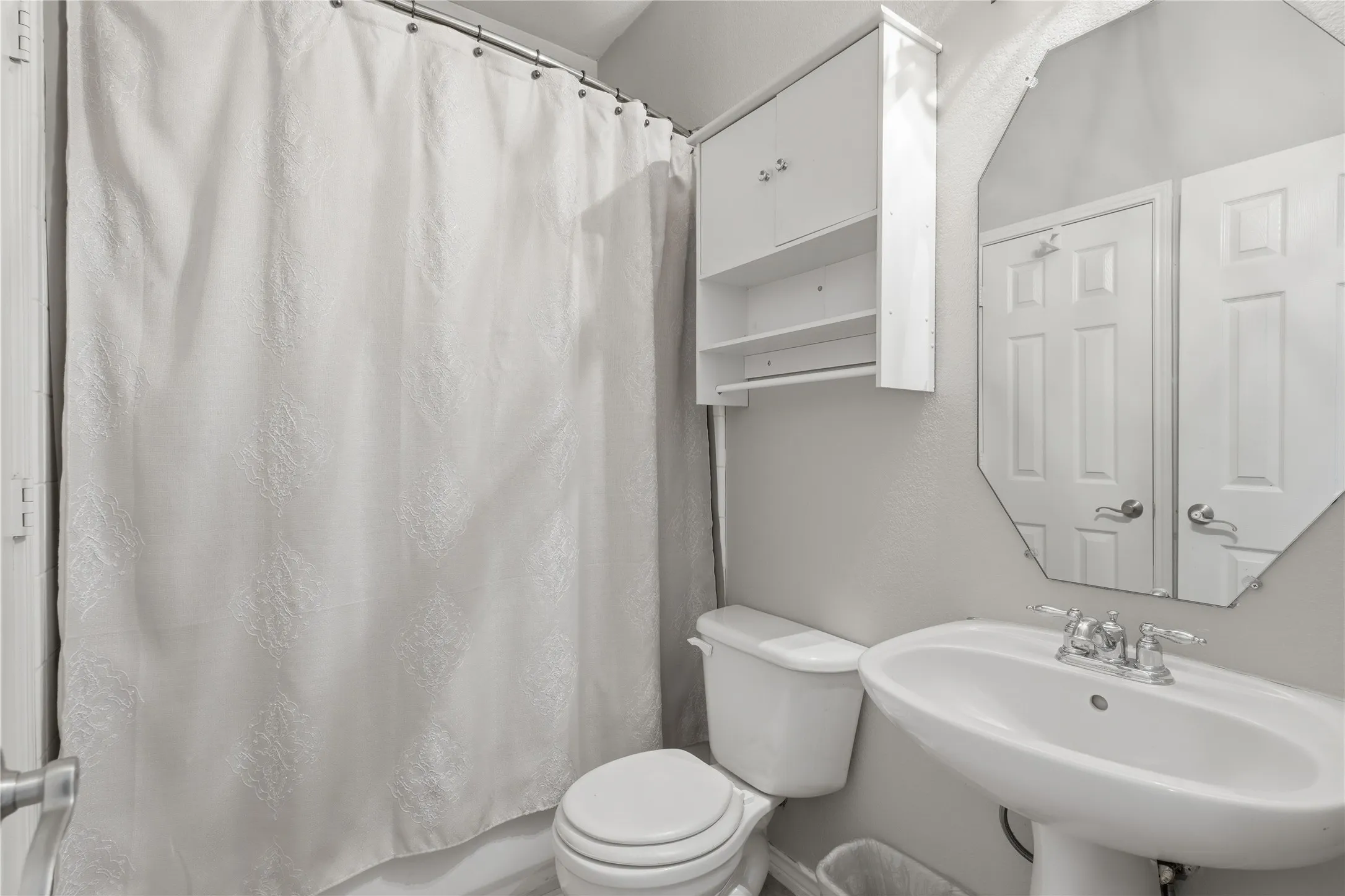 Bathroom featuring a pedestal sink with chrome faucet, octagonal wall mirror, white toilet, and built-in white wall cabinetry