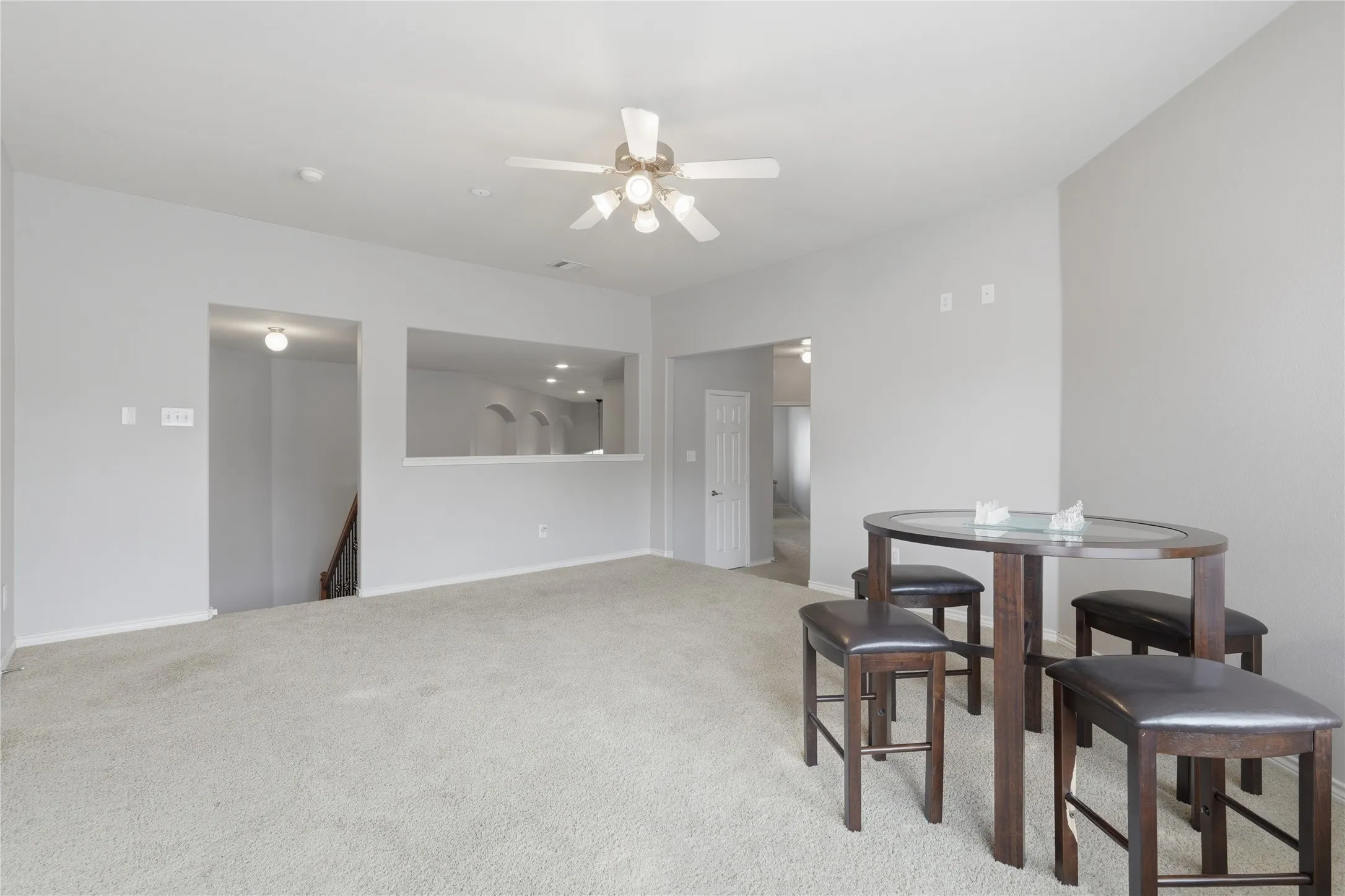 Carpeted room with light grey walls, featuring a ceiling fan with integrated lighting, a pass-through window with recessed lighting, and a staircase with wooden railings