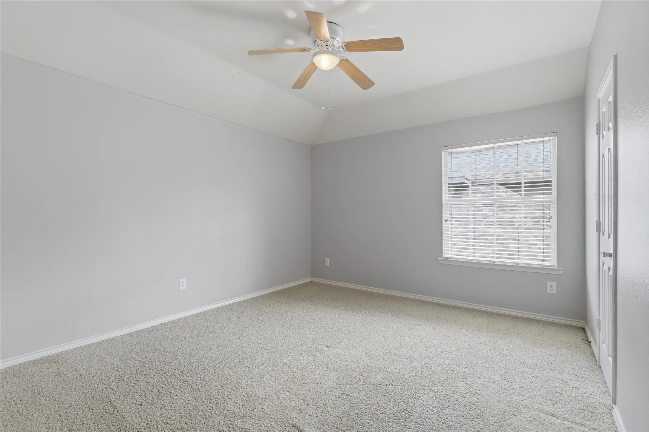 Spacious room featuring vaulted ceilings, light gray walls, plush carpeting, a multi-pane window with blinds, and a ceiling fan