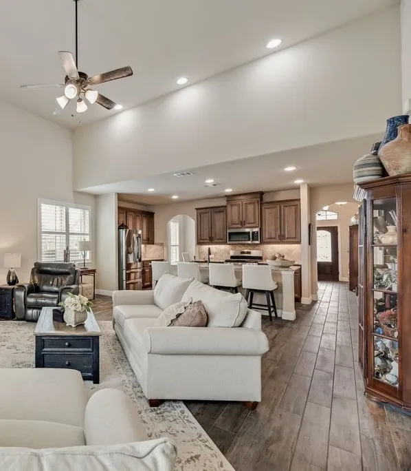 Partial View of the Area Labeled Cafe Area (Dining Area) on Builder Floor Plan from 2021. The Window Area across and behind the Sofa and Kitchen Island Chairs is the Cafe Area on the floor plan - measures approximately 17'9'' in length and 8' in Width. The photo includes partial views of Hall Entry, Kitchen, and Family Area/ Living Area on 1st level. Family Area in this photo shows a portion of the raised ceiling and ceiling fan with additional recessed lighting, and the Wood Look Ceramic Tile throughout first level.