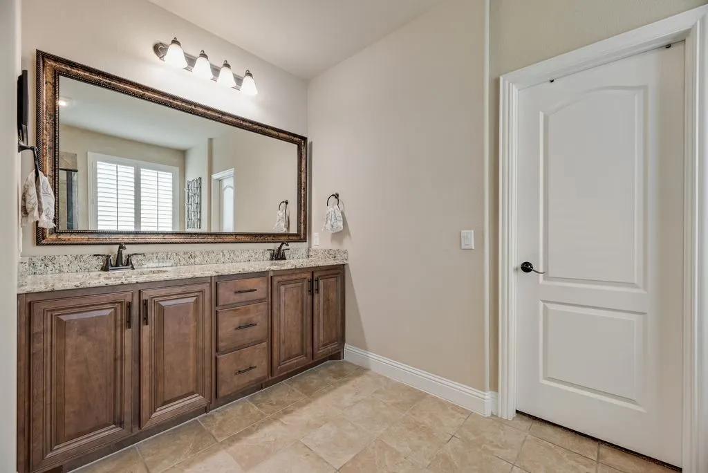 Partial View of Primary Bathroom - Upgraded Wood Cabinetry Finish on All Cabinets in the House - Nutmeg with the Black Glaze Finish - Double Vanity with Granite Counter tops, Framed Mirror, Cabinet Pulls added after Built in 2021. The closed door opens to the Large Walk In Primary Closet. No photo - schedule your visit to view tour the home to see onsite