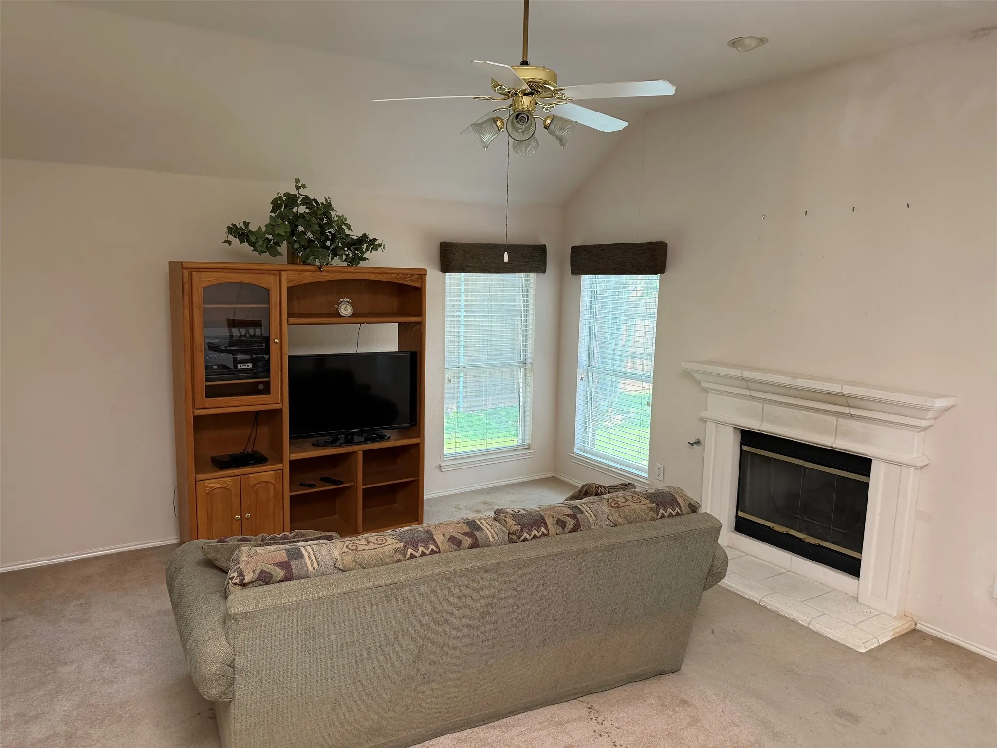 Spacious living area featuring a vaulted ceiling with a ceiling fan, a white tile-surround fireplace, and two windows with horizontal blinds