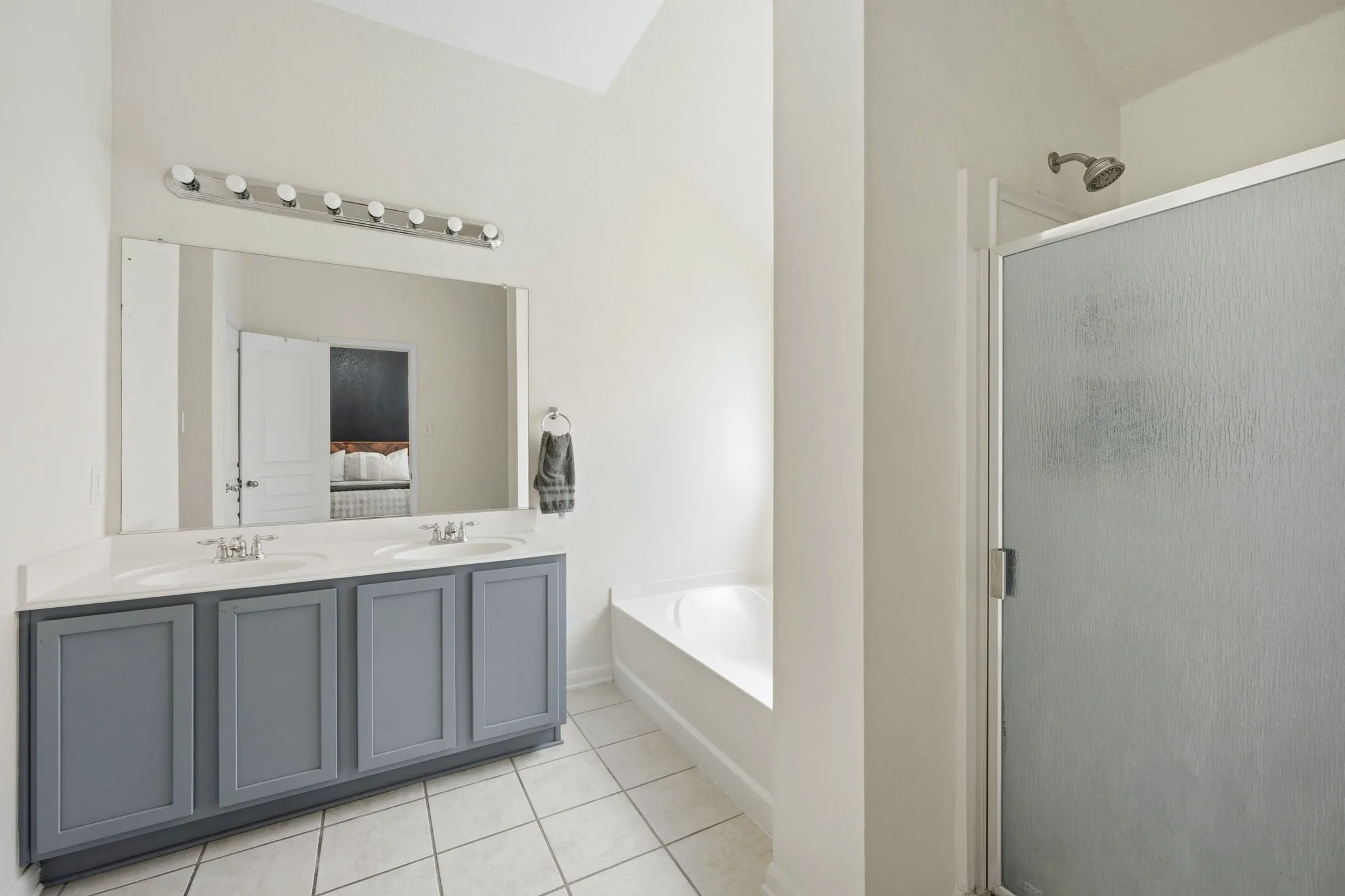 Bathroom featuring a double vanity with gray cabinetry and white countertops, a large mirror, a chrome light fixture, a built-in bathtub, and a separate shower with a frosted glass enclosure