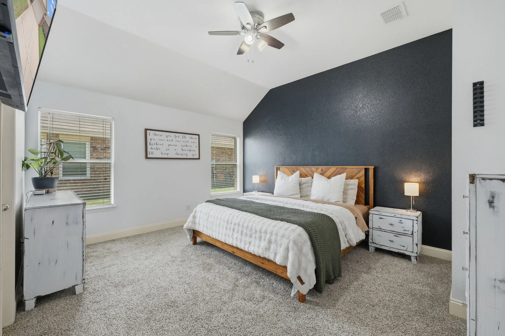 Vaulted ceiling bedroom featuring a dark accent wall, two windows with blinds, a ceiling fan, and grey carpet flooring