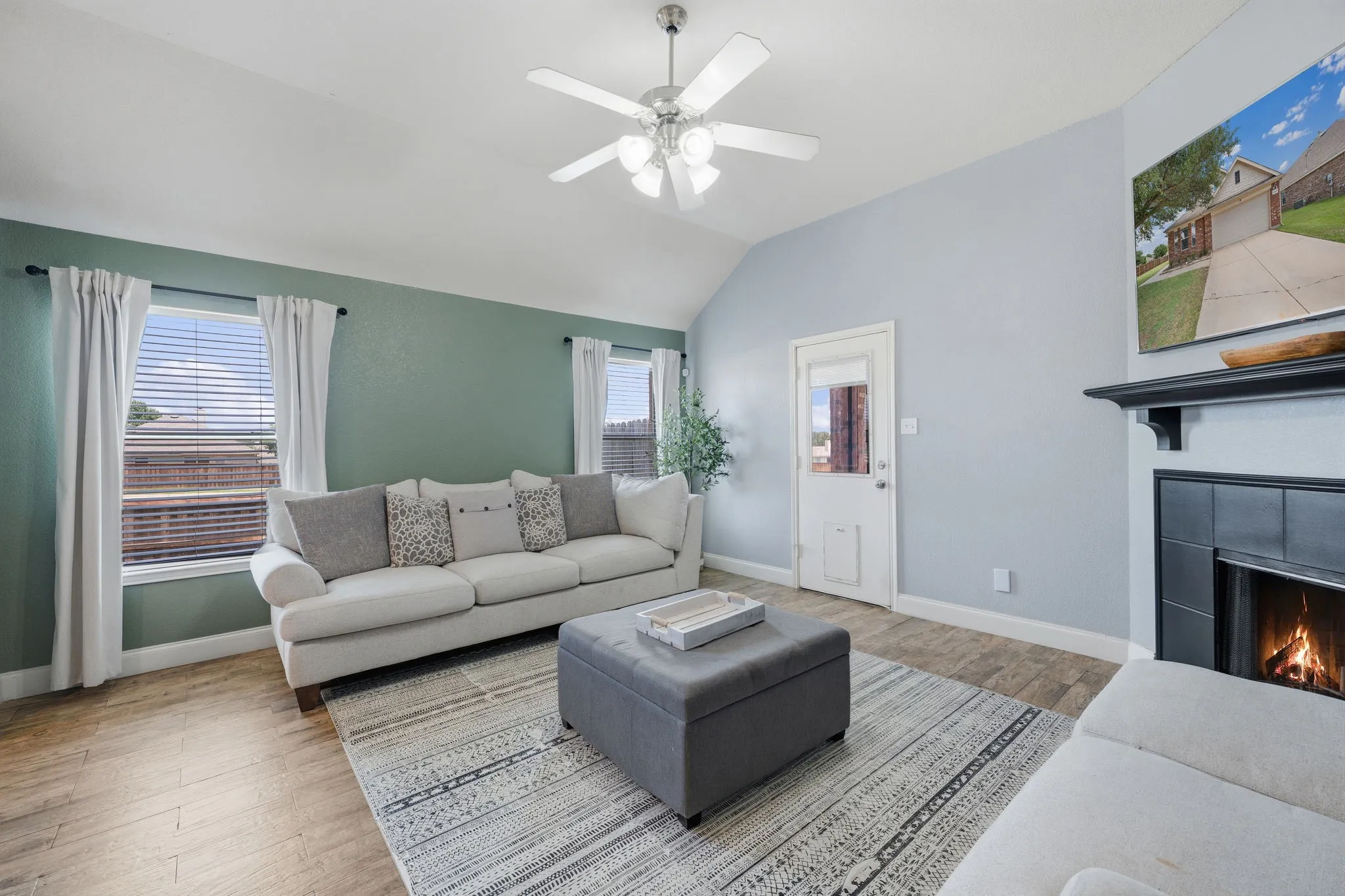 Living area featuring vaulted ceilings, wood-finish flooring, a ceiling fan with integrated lighting, a fireplace with a black tile surround and a dark wood mantle, and multiple windows