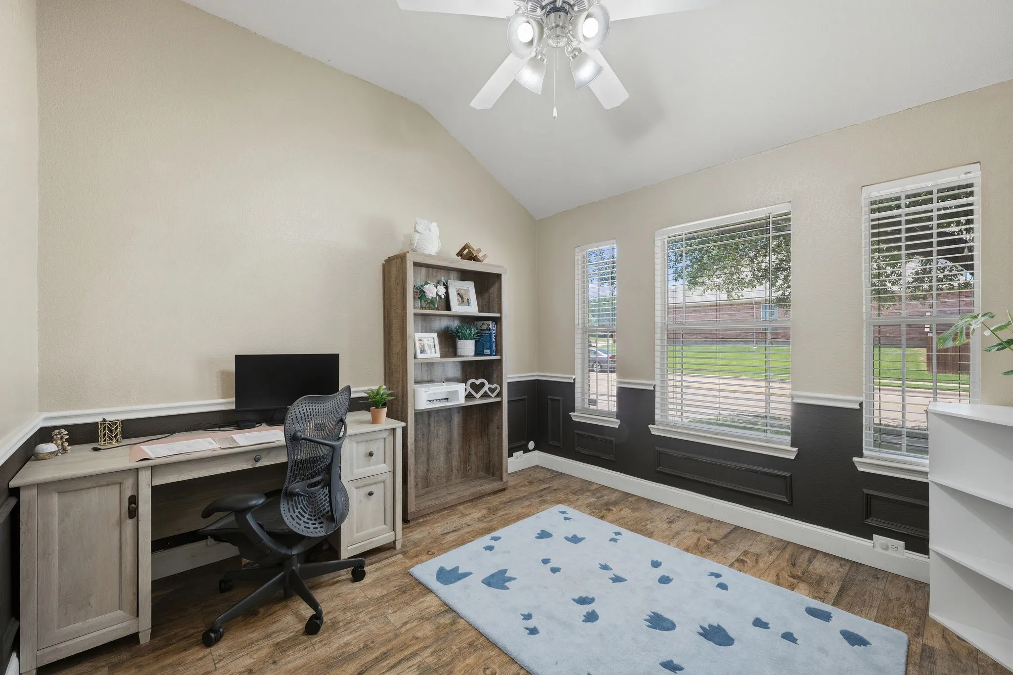 Vaulted ceiling room featuring wood-finish flooring, three windows with white blinds, and two-tone walls with dark wainscoting