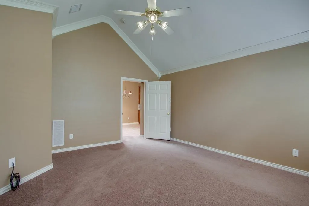 Spacious room featuring vaulted ceilings, a ceiling fan with integrated lighting, crown molding, an entry door, and carpet flooring