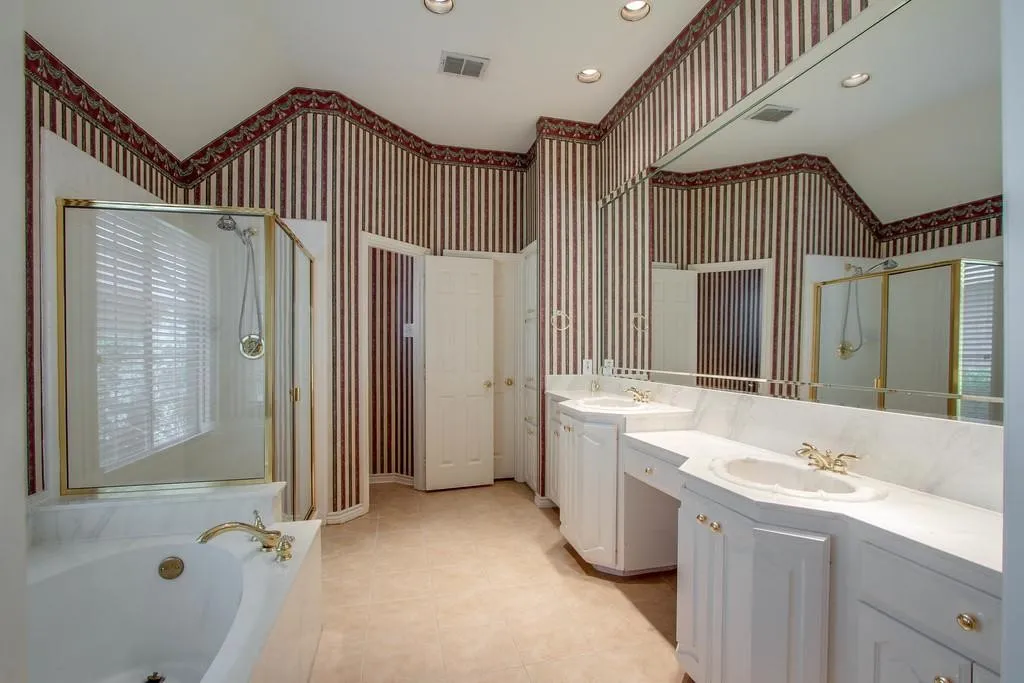 Full bathroom featuring dual vanity sinks set in white cabinetry with a marble-look countertop, a large built-in mirror, a built-in jetted tub, and a glass-enclosed shower with gold-tone fixtures