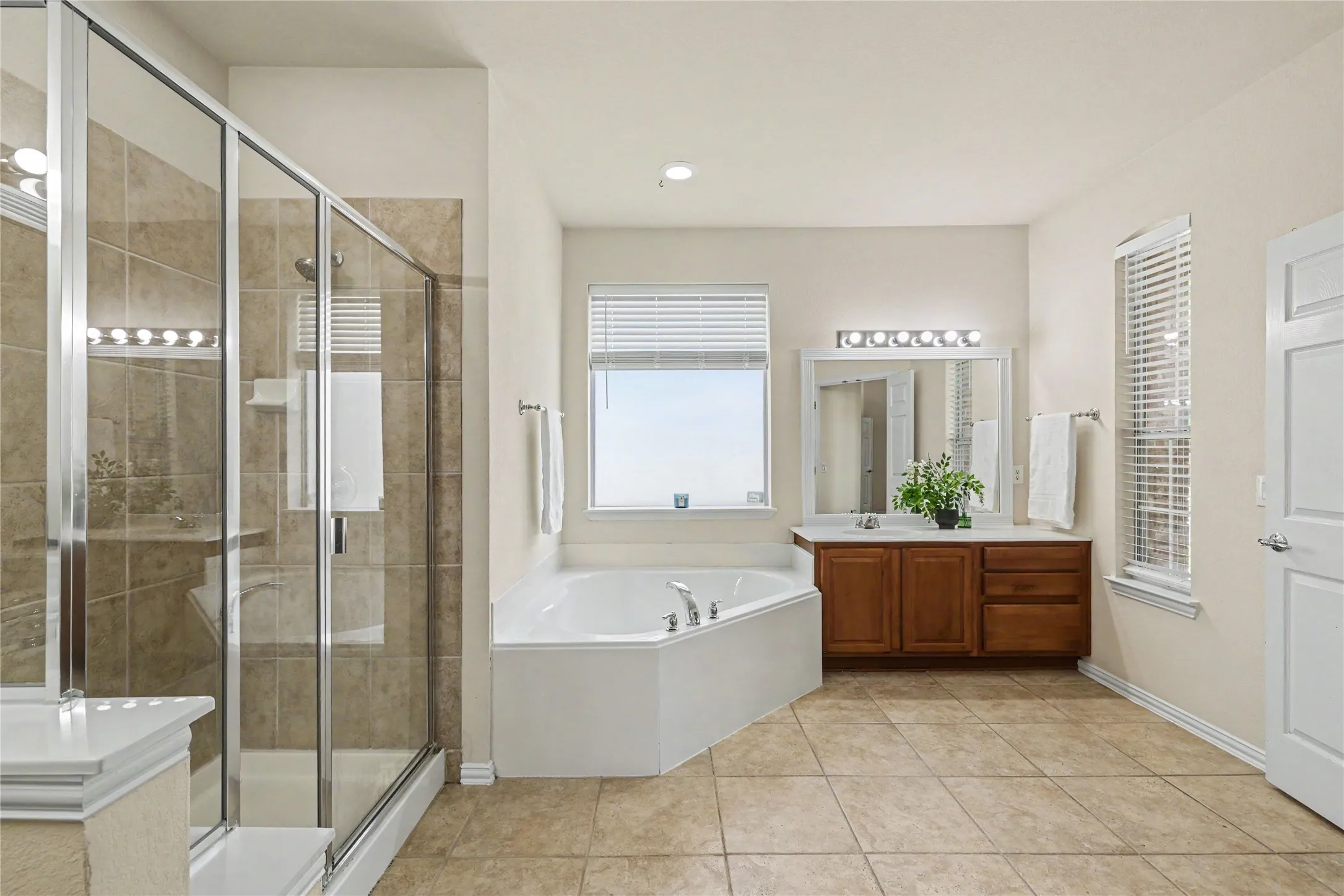 Spacious bathroom featuring an enclosed glass shower, a corner soaking tub, a double vanity with wood cabinetry, and large format tile flooring