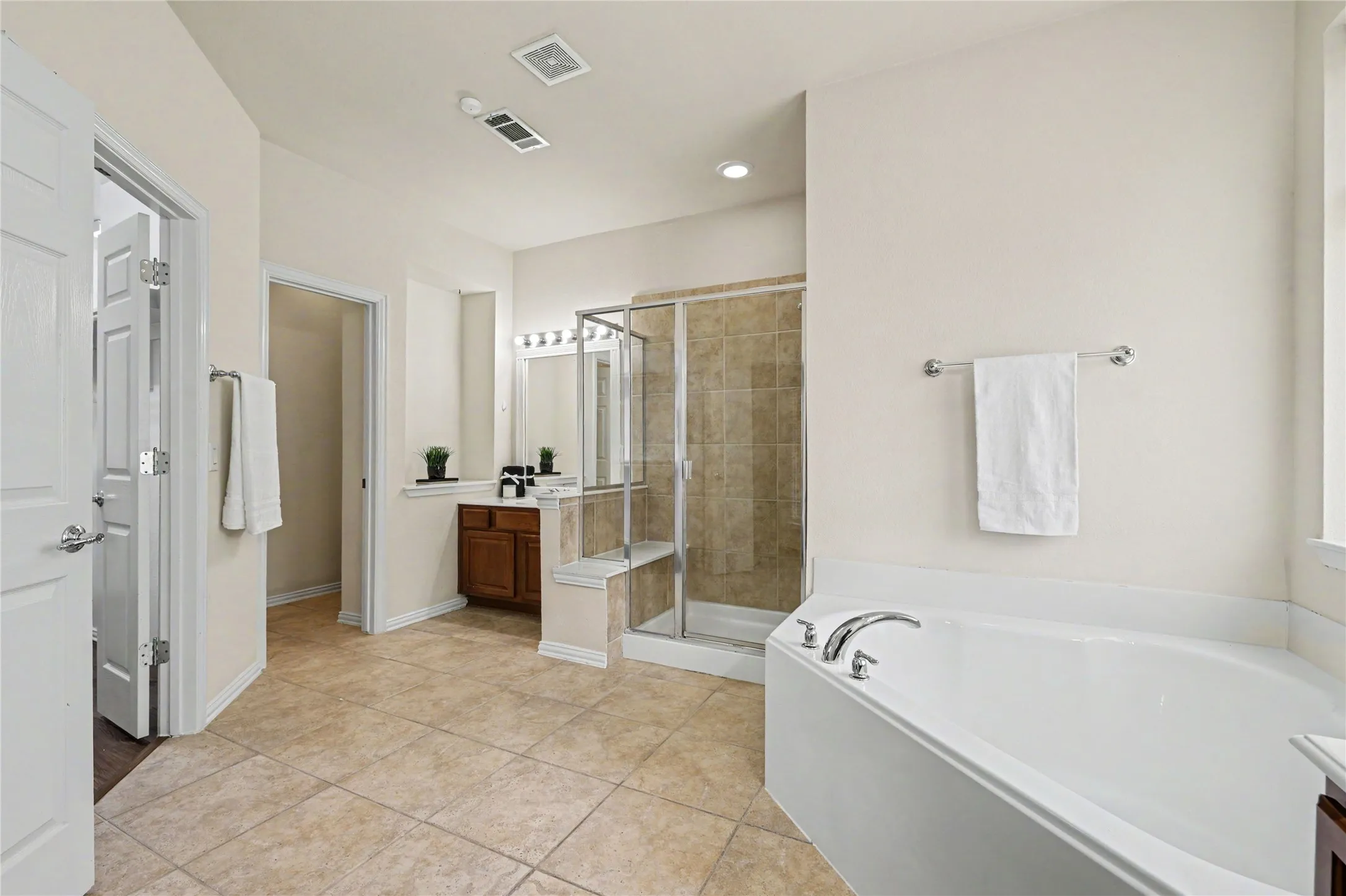 Bathroom featuring a corner soaking tub with chrome fixtures, a glass-enclosed shower with tiled walls, a wood-finish vanity with a countertop, and large format tile flooring