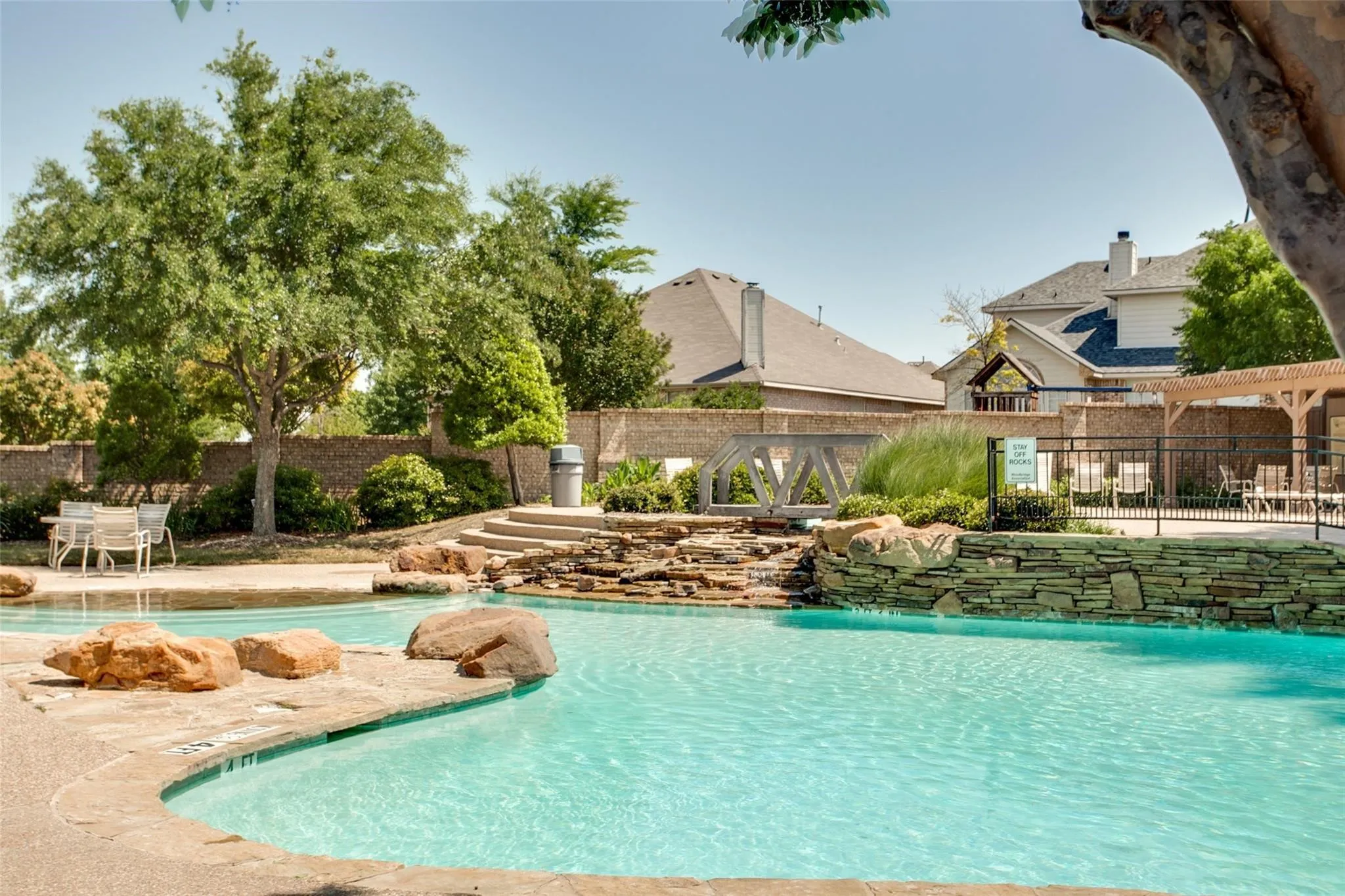 Community swimming pool with rock features and a waterfall, concrete decking, and an adjacent patio area with a pergola