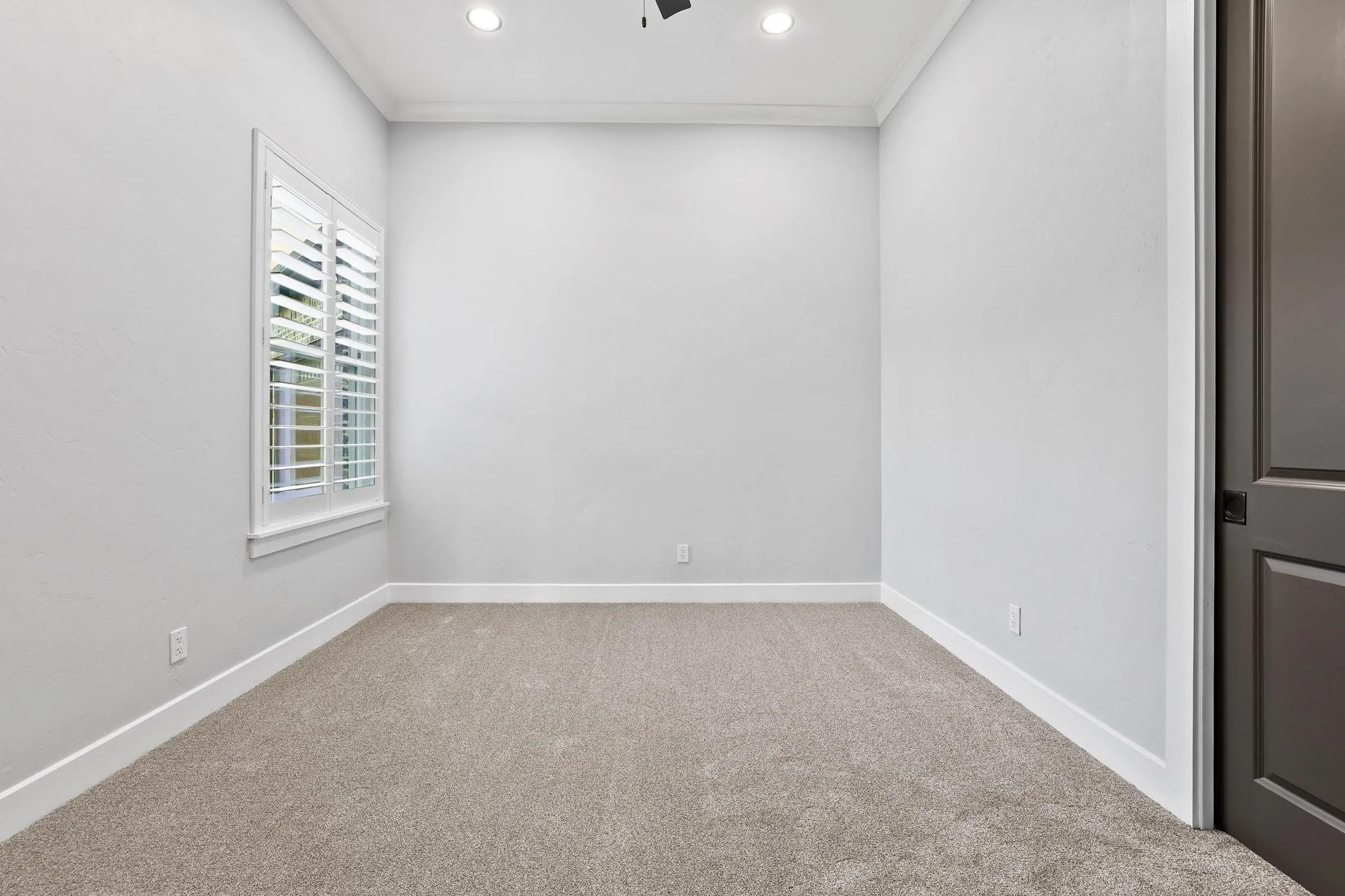 Bright room with neutral wall color, plush carpet, recessed lighting, and a window featuring white plantation shutters