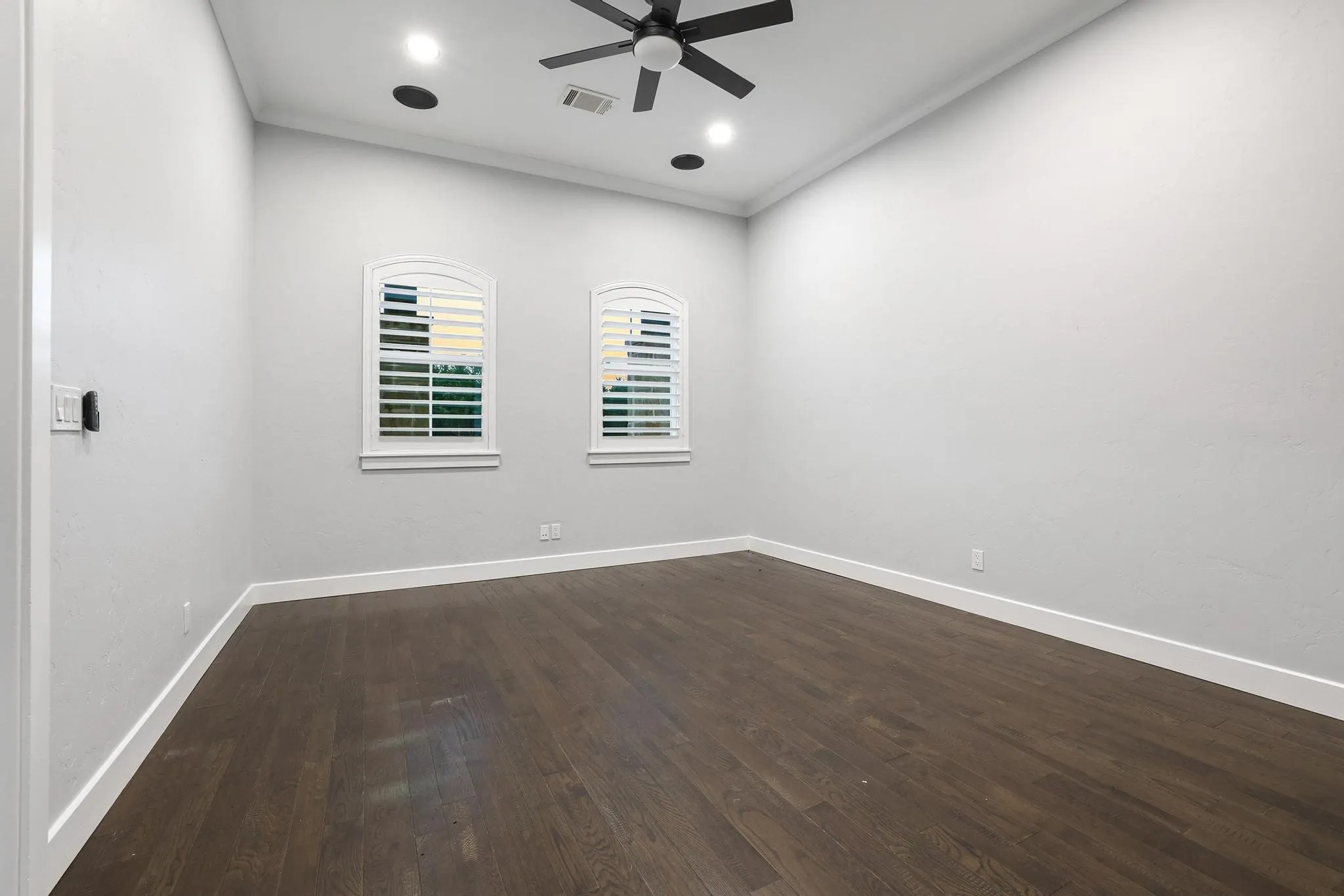 Spacious room featuring wood-finish flooring, two arched windows with plantation shutters, a ceiling fan, recessed lighting, and crown molding