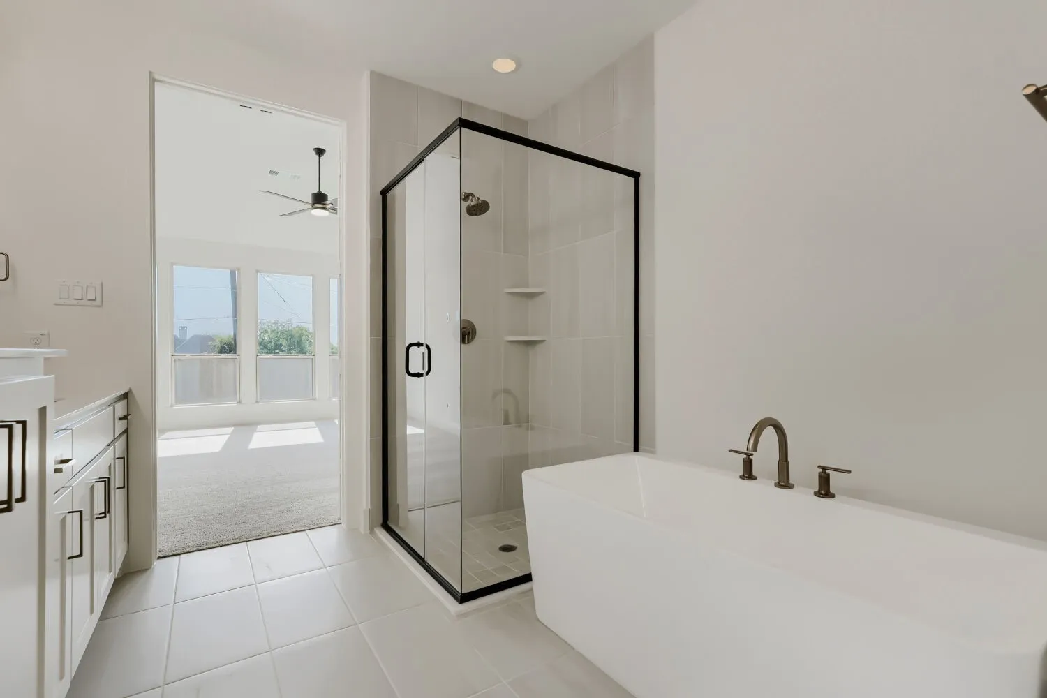 Freestanding soaking tub with dark-bronze fixtures, glass-enclosed shower with dark-bronze frame, white tiled flooring, and light-toned vanity with dark hardware