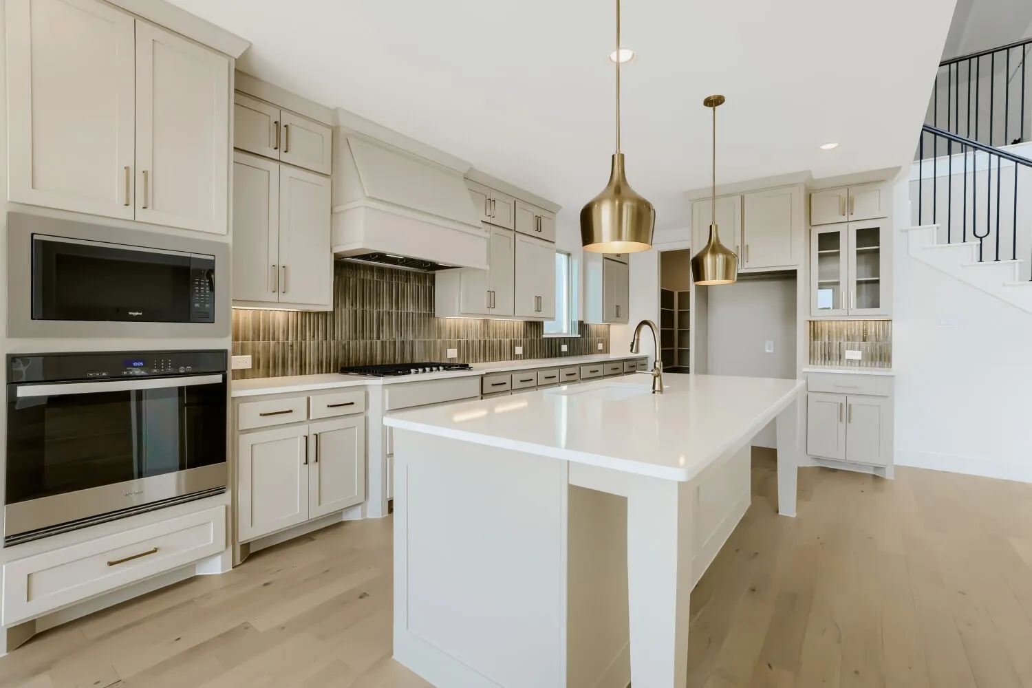 Gourmet kitchen featuring white cabinetry with glass-front upper cabinets, stainless steel built-in appliances including a wall-mounted oven, quartz countertops, a large waterfall-edge island with seating, brass pendant lighting, and wood-finish flooring throughout the open-concept space