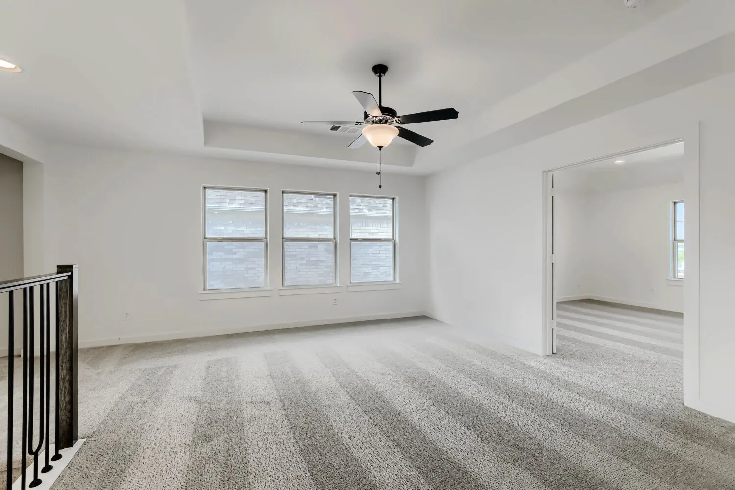 Spacious open room featuring striped carpet flooring, abundant natural light from a trio of large multi-pane windows, black ceiling fan with integrated lighting, and seamless access to an adjoining bright interior space