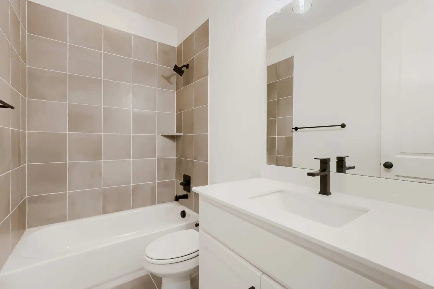 Full bathroom featuring a white tub, light-toned tiled shower surround, recessed shower shelf, white vanity with integrated sink, and matte black fixtures