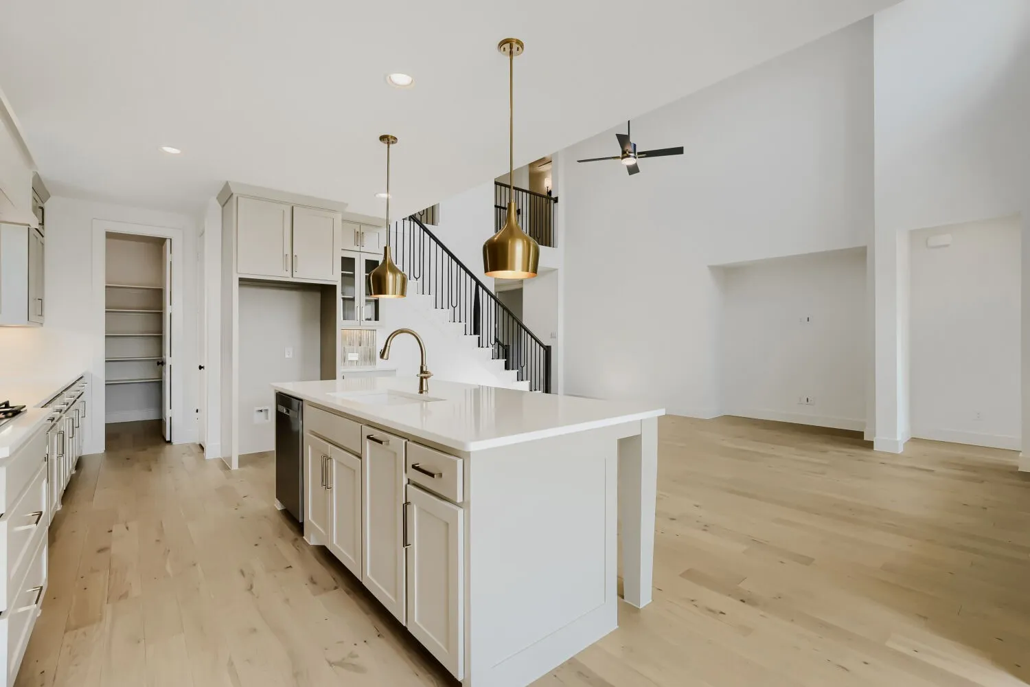 Open-concept kitchen and living area featuring a large waterfall-edge island with white cabinetry, stainless steel sink with gooseneck faucet, wide-plank wood-finish flooring throughout, pendant lighting with brass and black metal fixtures, and soaring ceilings with recessed lighting
