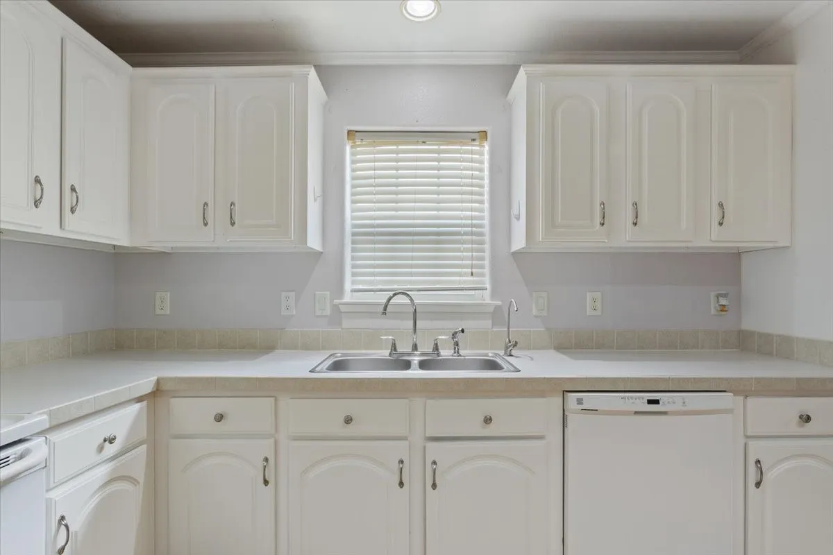 Kitchen featuring white cabinetry, light-toned countertops, a stainless steel double basin sink with a gooseneck faucet, and a white built-in dishwasher