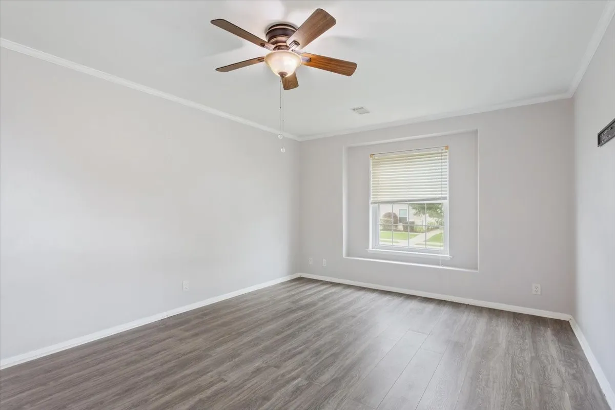 Spacious room featuring wood-finish flooring, white baseboards, crown molding, a window with blinds, and a ceiling fan with light fixture