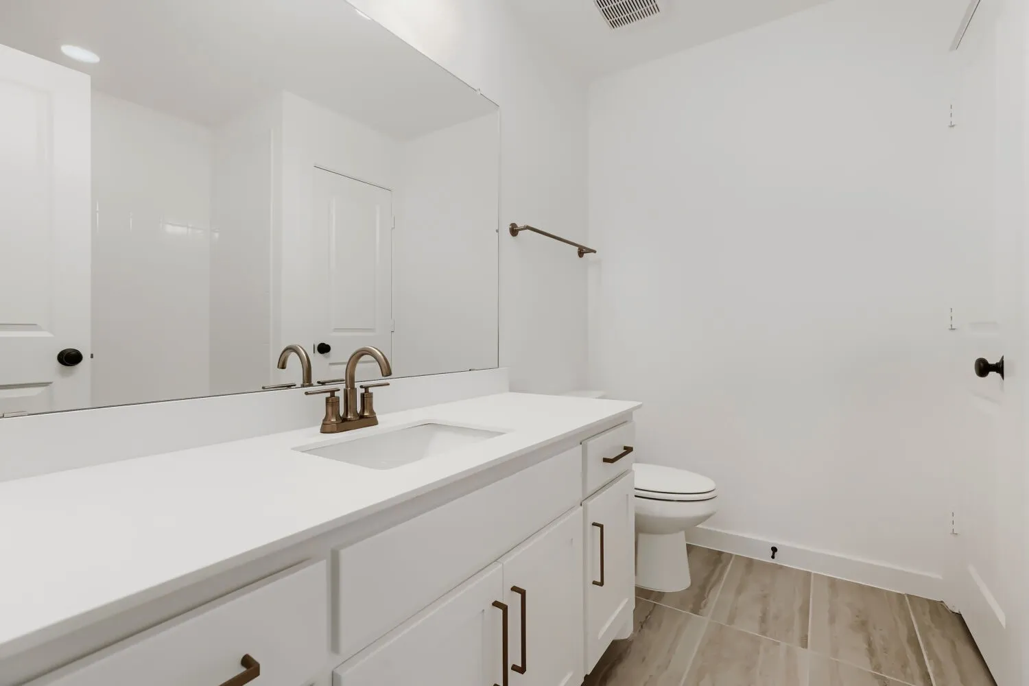 White vanity with integrated sink, white countertop, bronze-finish faucet, elongated mirror, and neutral-tone tile flooring