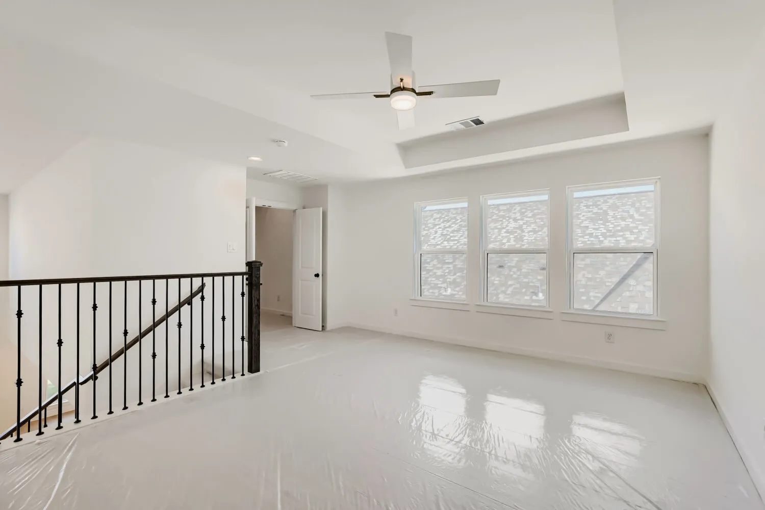 Spacious room featuring three windows, a ceiling fan with integrated lighting, and a black wrought iron staircase railing