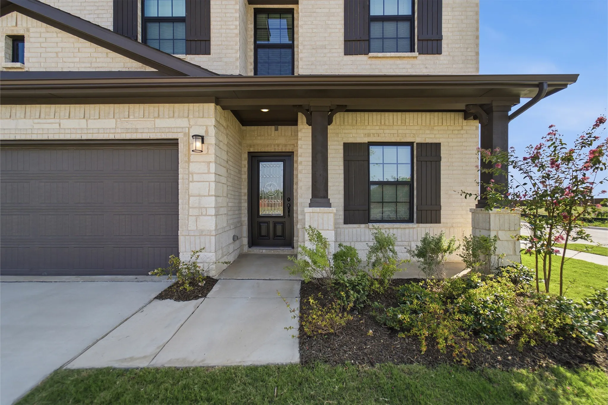 Cream-colored brick exterior featuring dark brown window shutters, a covered entry with decorative columns, a dark panel garage door, and a glass-paneled front door