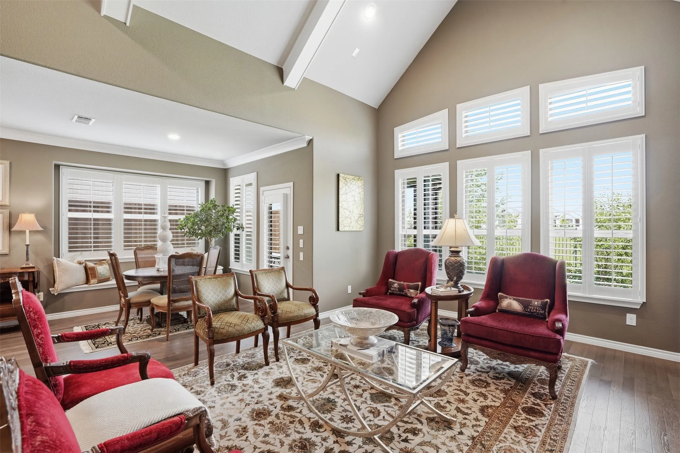 Spacious living area featuring a vaulted ceiling with exposed beams, wood-finish flooring, and multiple windows with plantation shutters