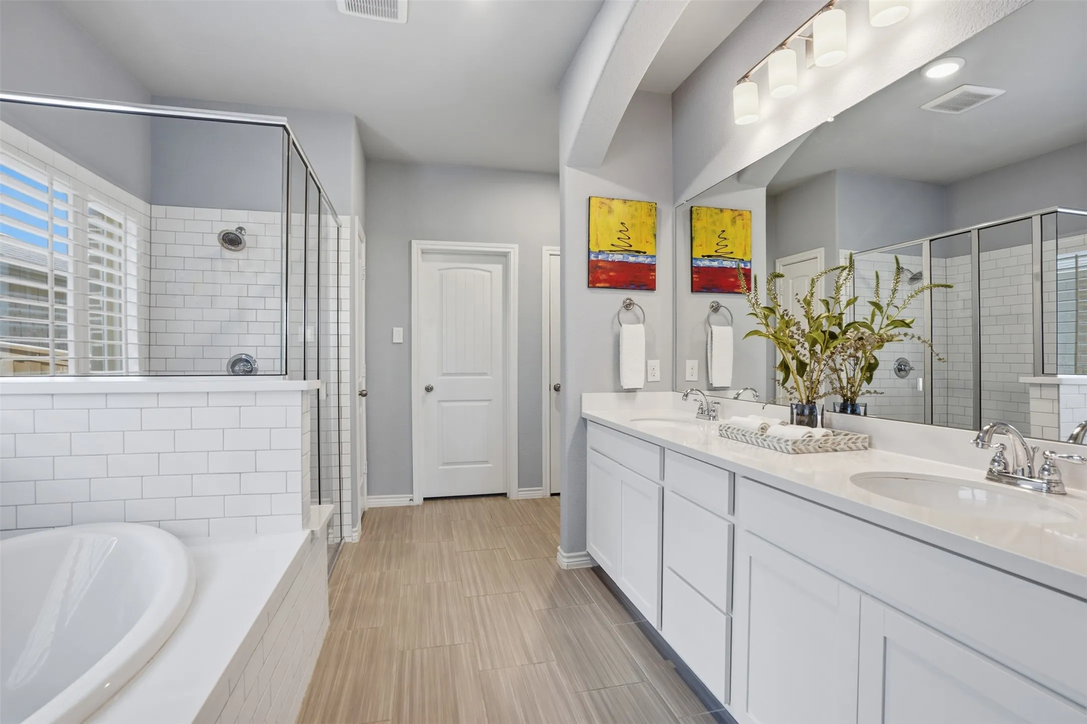 Spacious bathroom featuring a dual vanity with white cabinetry and light-toned countertops, a large mirror with overhead lighting, a built-in tub, and a shower with white subway tile and glass enclosure