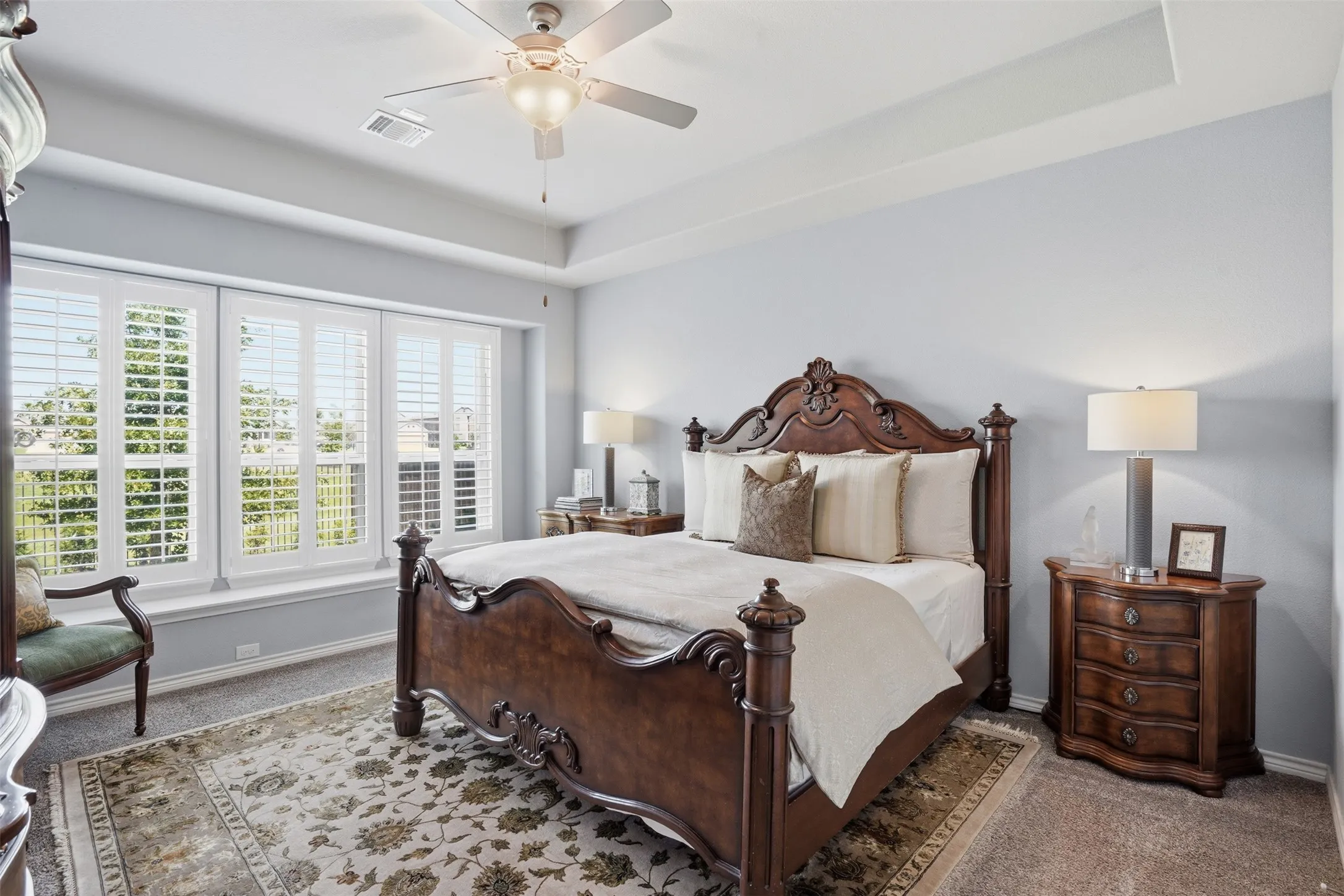 Bedroom featuring a tray ceiling with a ceiling fan, three large windows with white plantation shutters, light gray wall paint, and carpet flooring