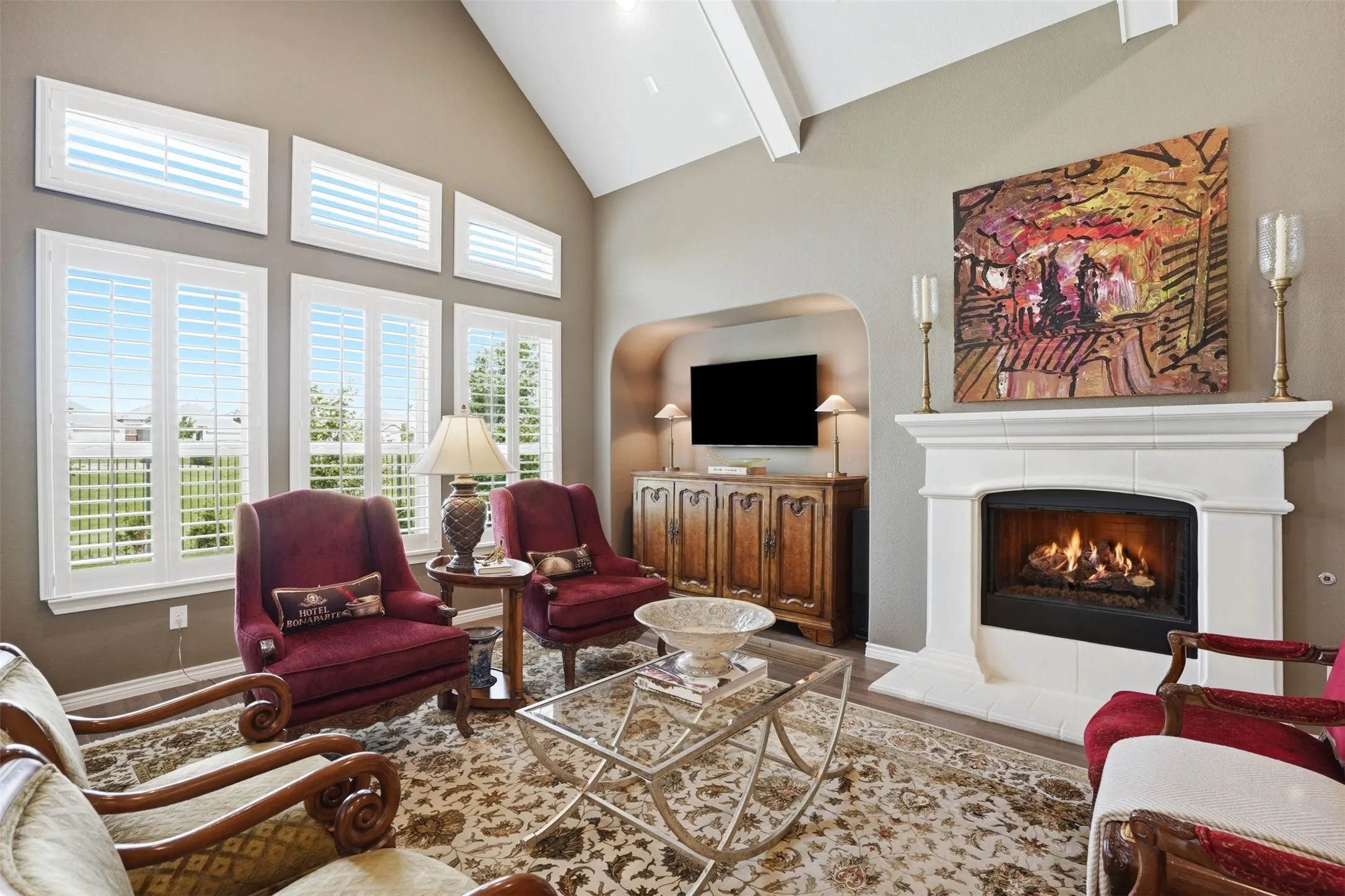 Living area featuring a vaulted ceiling with exposed beams, plantation shutters, a decorative fireplace with a mantel, an arched alcove, and a patterned area rug