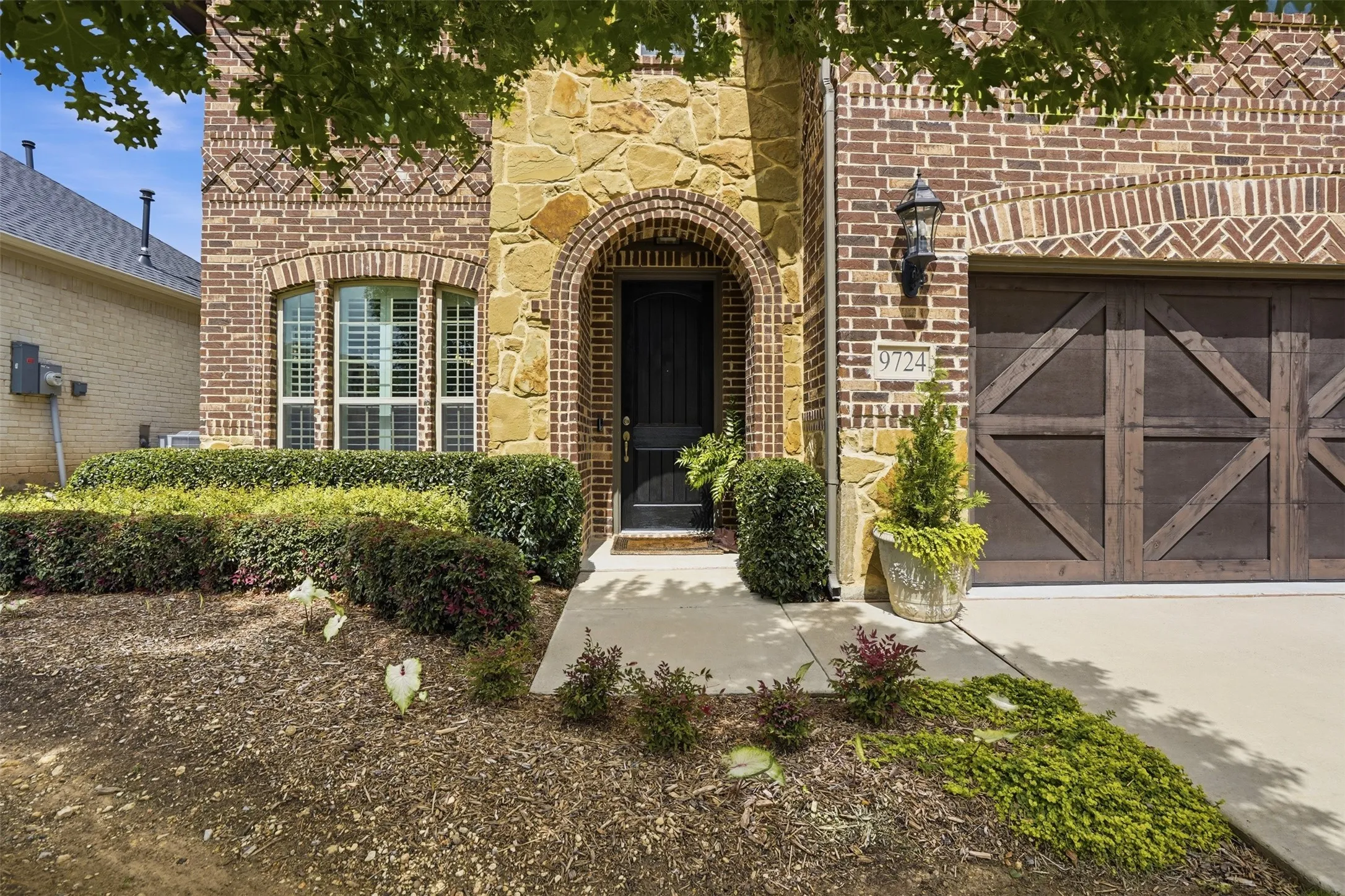 Brick and stone exterior featuring an arched entryway, decorative garage door, and mature landscaping