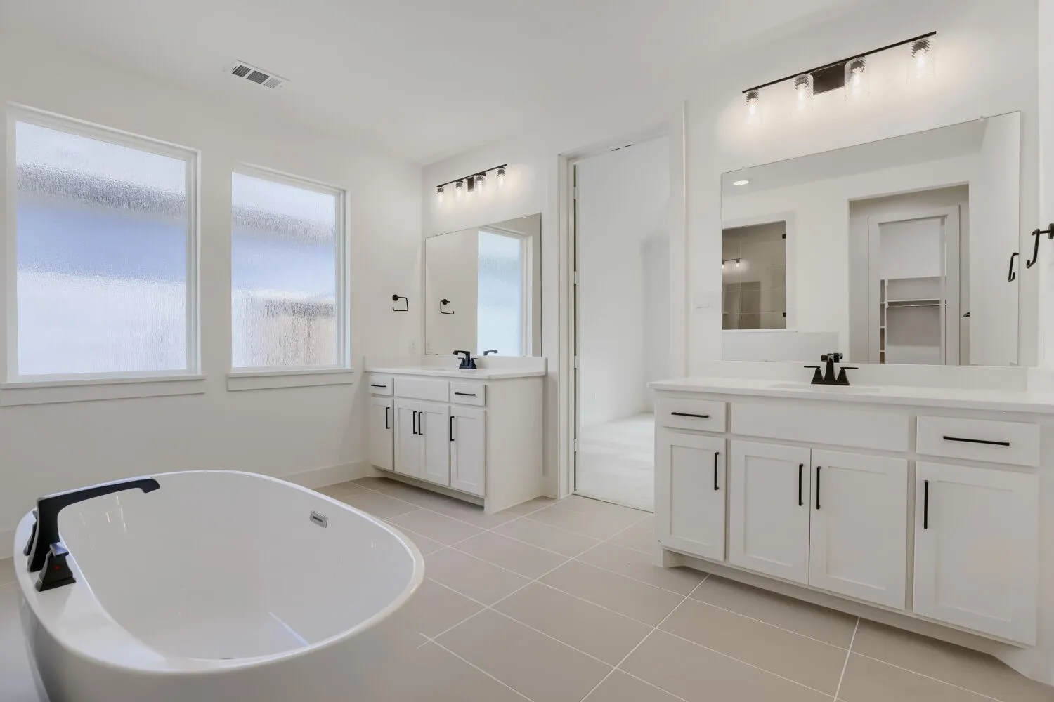 Freestanding soaking tub with matte black fixtures, dual vanity with shaker-style cabinetry, large mirror, and tiled flooring
