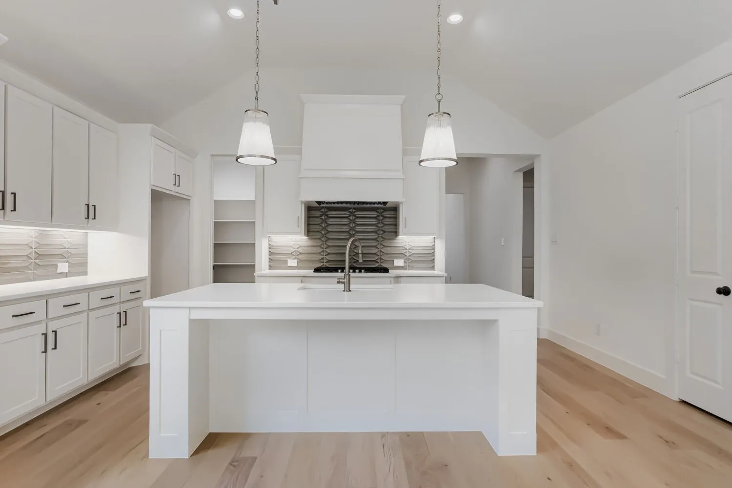 Chef's kitchen featuring a large center island with waterfall-edge countertop, white shaker-style cabinetry throughout, stainless steel sink with pull-down faucet, pendant lighting, subway tile backsplash, and wood-finish flooring