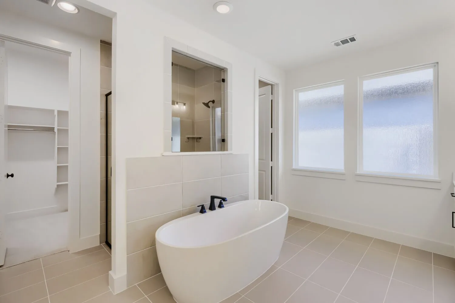 Freestanding soaking tub with matte black fixtures, tile flooring, glass-enclosed shower, two privacy windows, and recessed lighting