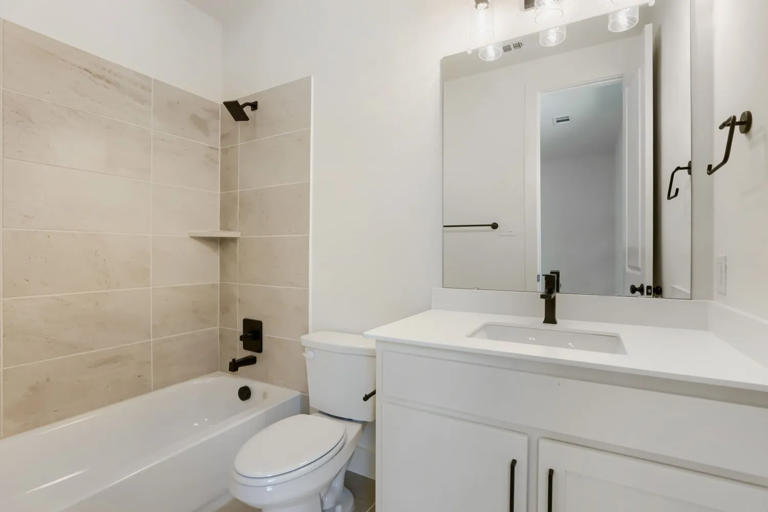 Contemporary bathroom featuring a white vanity with integrated sink and ample countertop space, large framed mirror with integrated lighting, matte black hardware accents, ceramic tile shower surround with built-in shelving, and a soaking tub with chrome fixtures