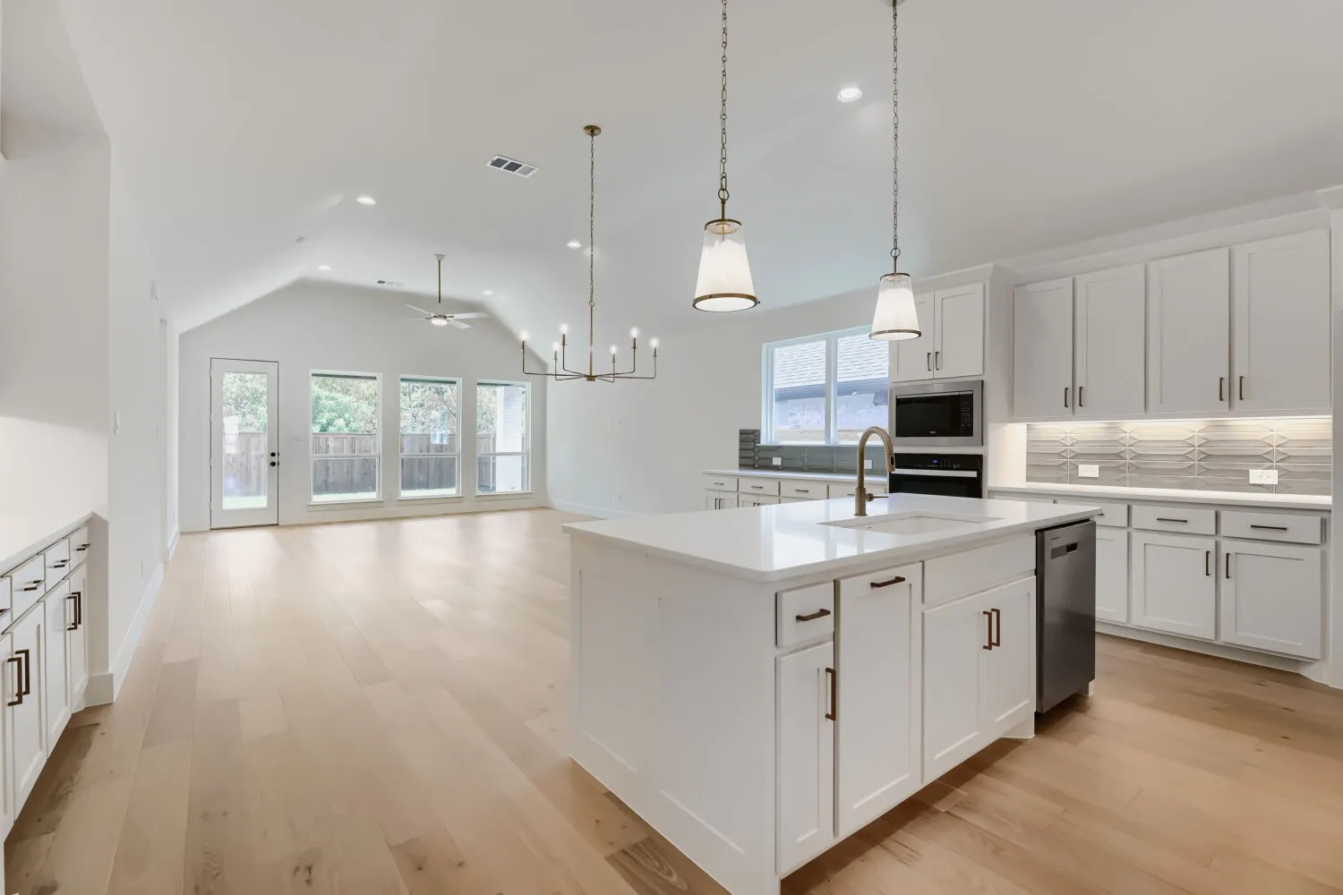 Open-concept living space with vaulted ceilings, light wood-finish flooring, white kitchen cabinetry, a large kitchen island with a sink, and a built-in microwave and oven combination