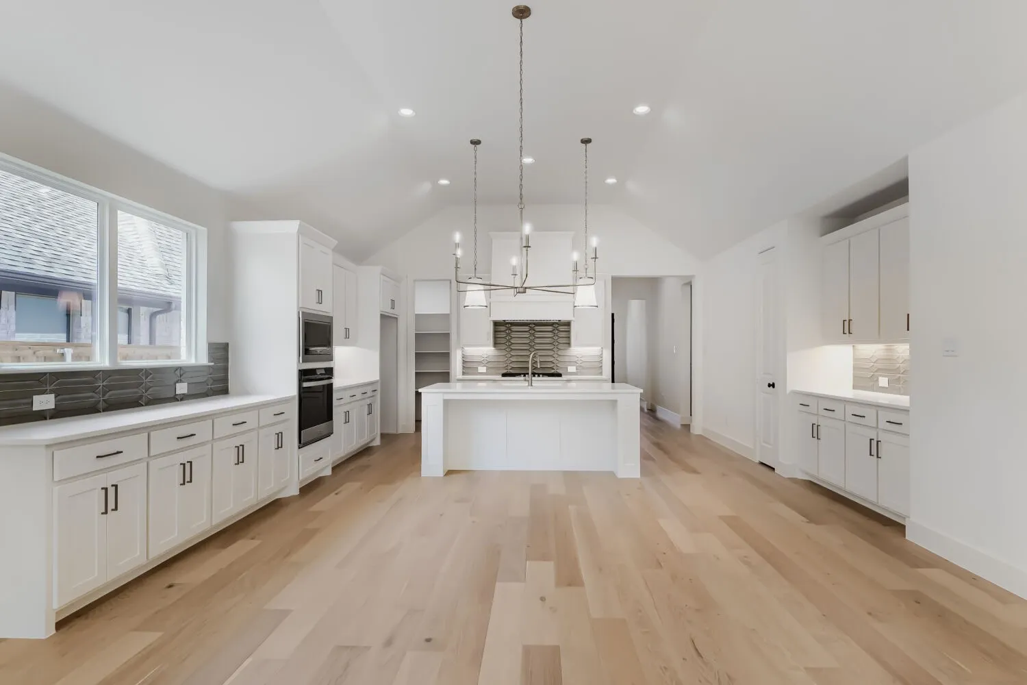 Expansive open-concept kitchen and dining area featuring white cabinetry, stainless steel appliances, a large center island with waterfall edge, wide-plank wood-finish flooring, and a multi-pendant light fixture suspended above the island