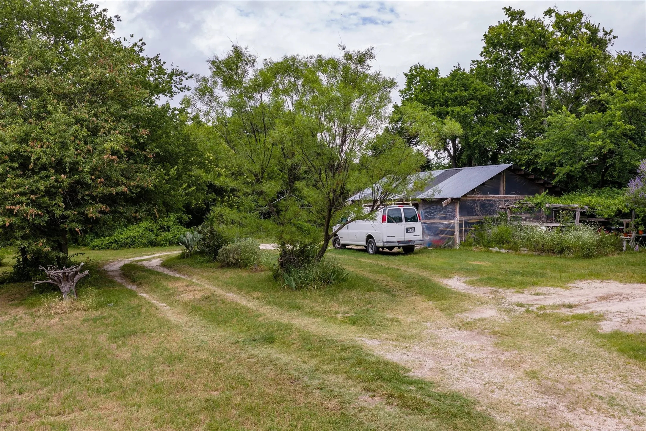 Gravel driveway leading to a metal-roofed outbuilding with fencing, surrounded by mature trees and natural landscaping