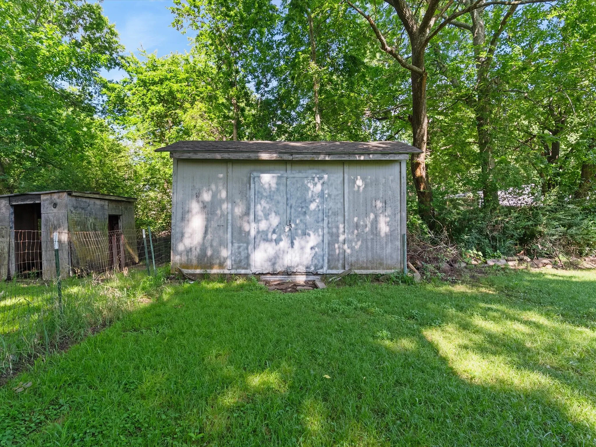 Storage shed with a pitched roof and double doors, accompanied by a smaller outbuilding, both surrounded by mature trees and lush greenery