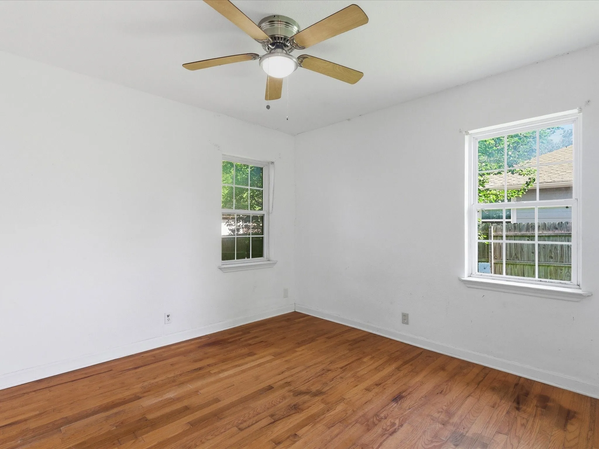 Wood-finish flooring, white painted walls, a ceiling fan with light fixture, and two window openings with white trim