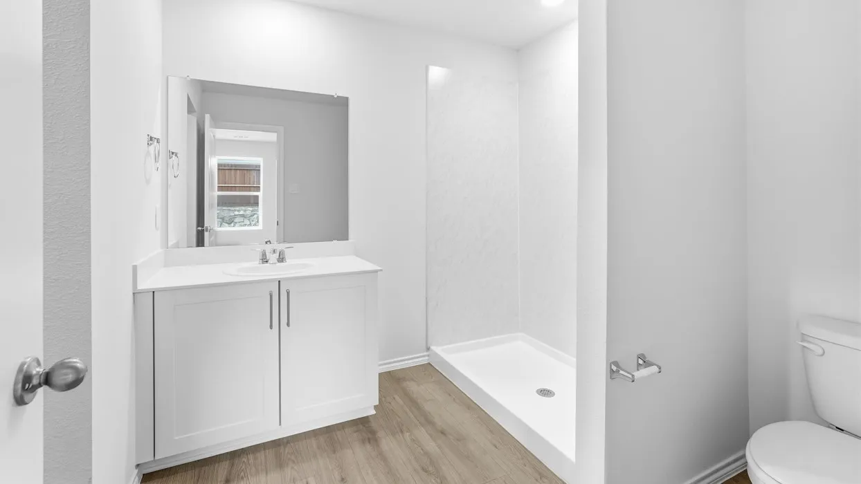 Bright bathroom featuring a white single-sink vanity with a light-toned countertop, a rectangular mirror, wood-finish flooring, and a stand-up shower with a white base