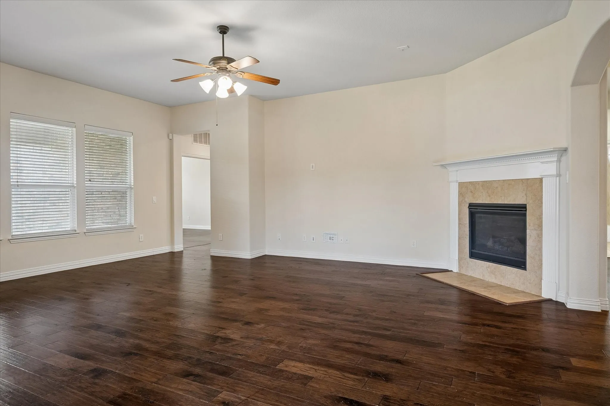 Spacious living area featuring rich wood-finish flooring, a contemporary fireplace with a stone surround, multiple windows with blinds, a ceiling fan with integrated lighting, and arched architectural details