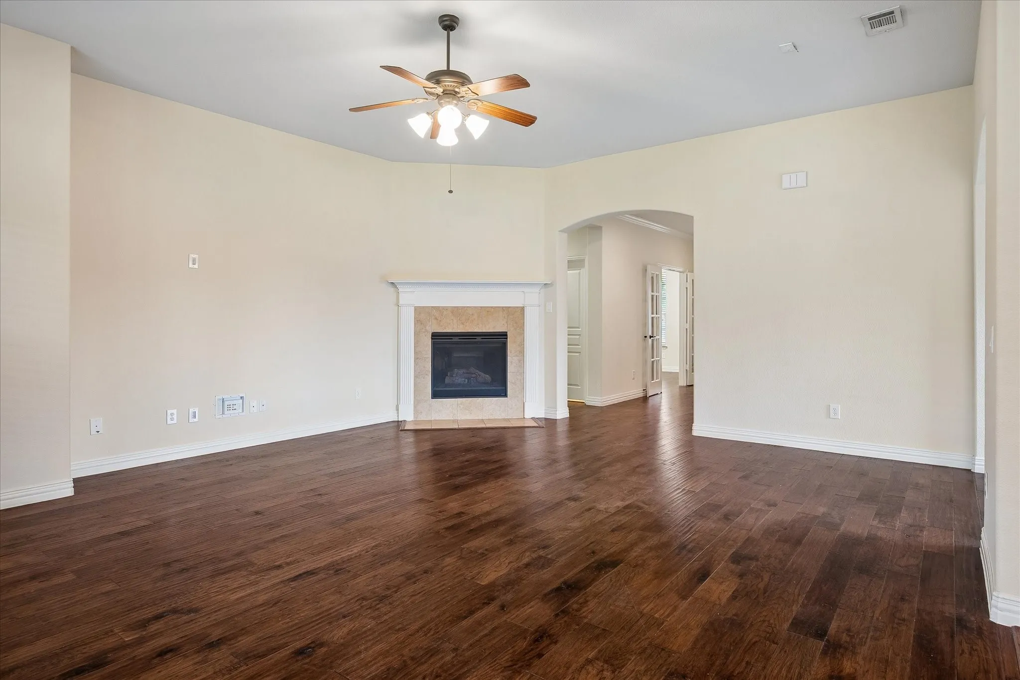 Wood-finish flooring, fireplace with stone surround and white mantel, arched doorway, ceiling fan with integrated lighting, and white baseboards