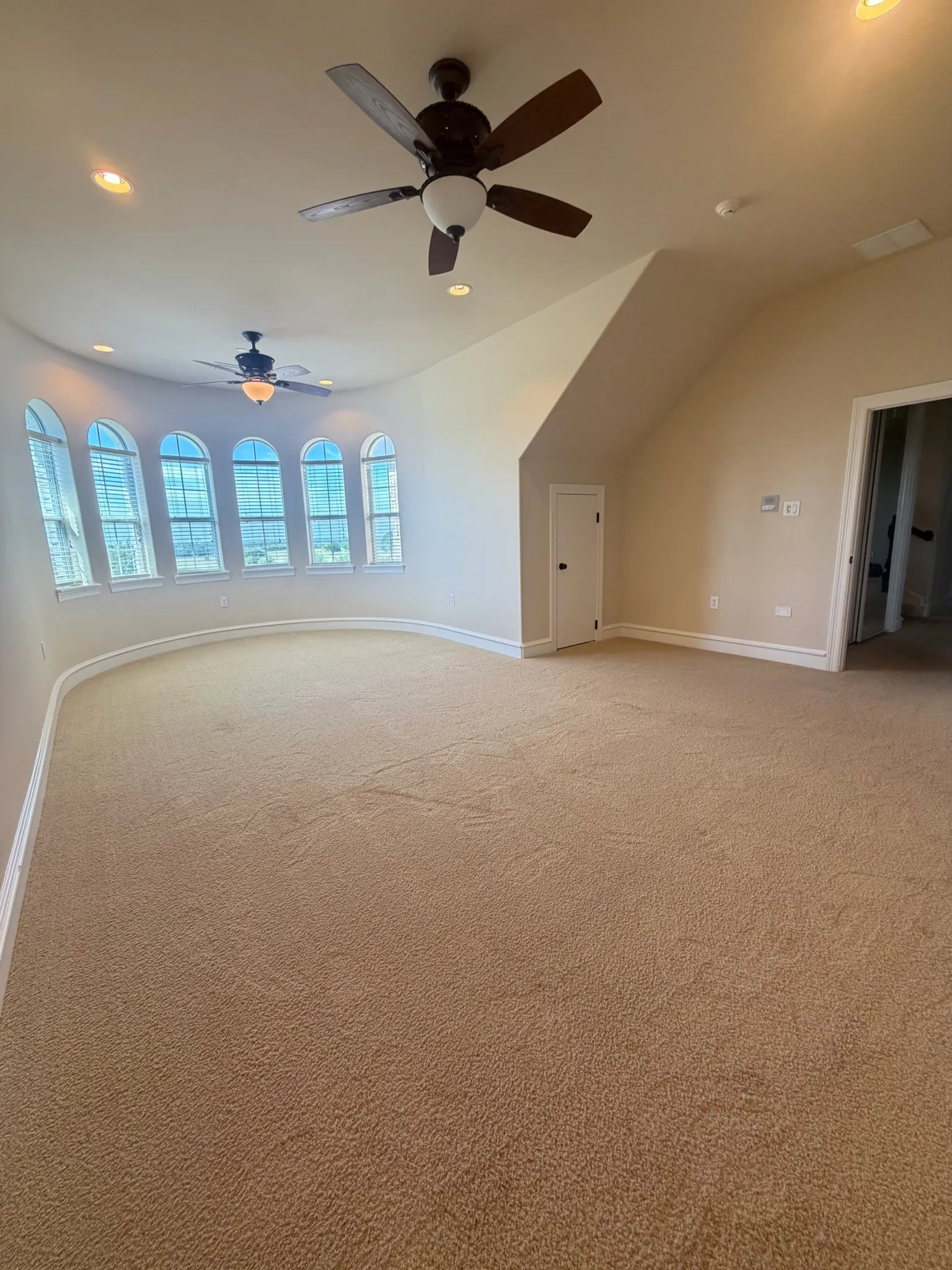 Spacious carpeted room featuring a curved wall of arched windows, two ceiling fans with integrated lighting, and recessed ceiling lights