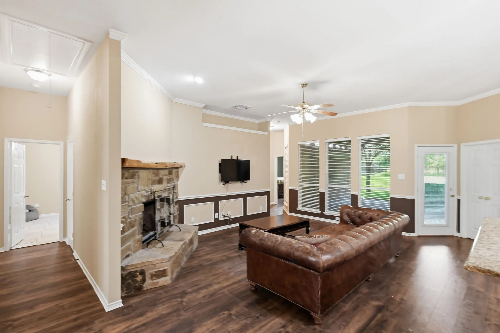 Spacious living area featuring wood-finish flooring, a stone fireplace with a substantial mantel, and multiple windows offering views of the exterior