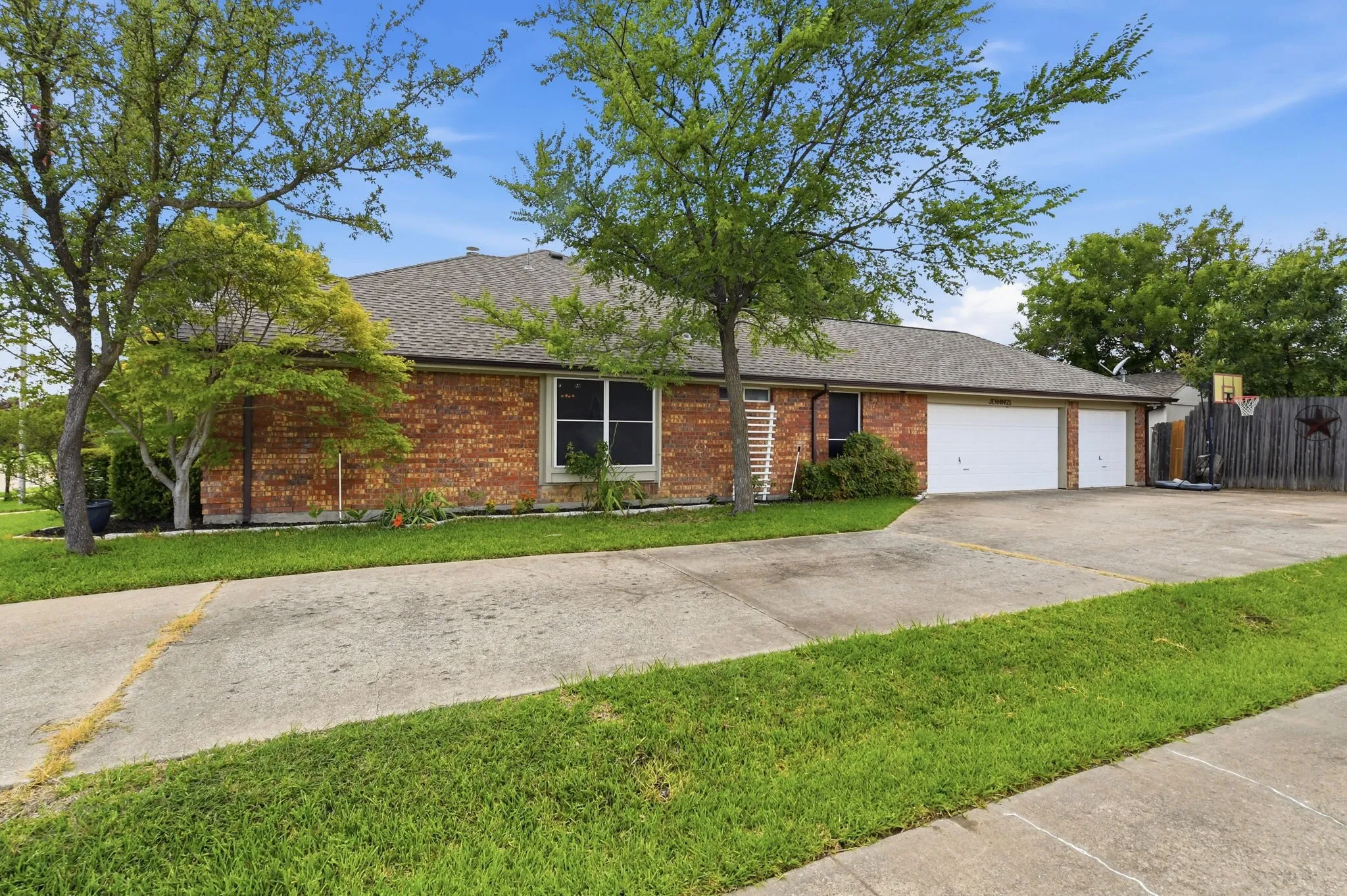 Brick exterior with a dark shingle roof, featuring mature trees, a well-maintained lawn, and a wide concrete driveway leading to a multi-car garage with white doors