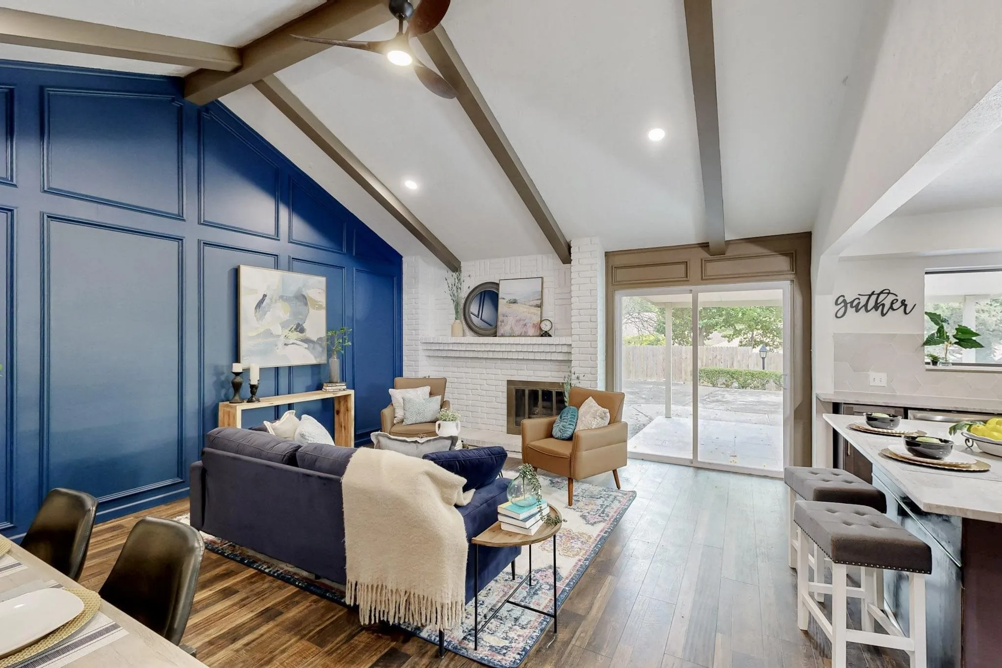 Vaulted ceiling with exposed wood beams, accent wall with panel molding, wood-finish flooring, brick fireplace surround, and sliding glass doors to the exterior patio
