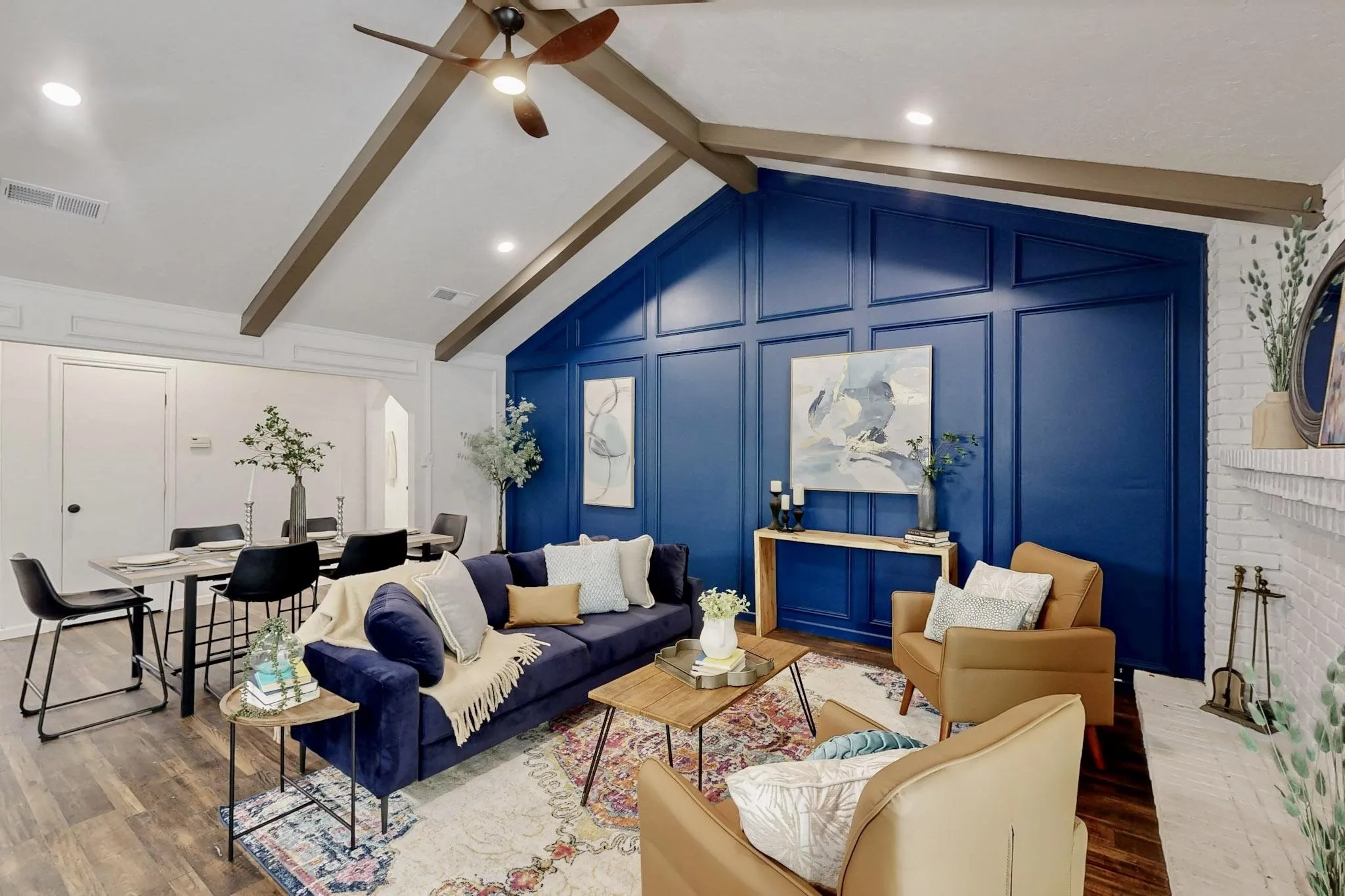 Vaulted ceiling with exposed wood beams, accent wall with panel molding, wood-finish flooring, white painted brick fireplace, and recessed lighting
