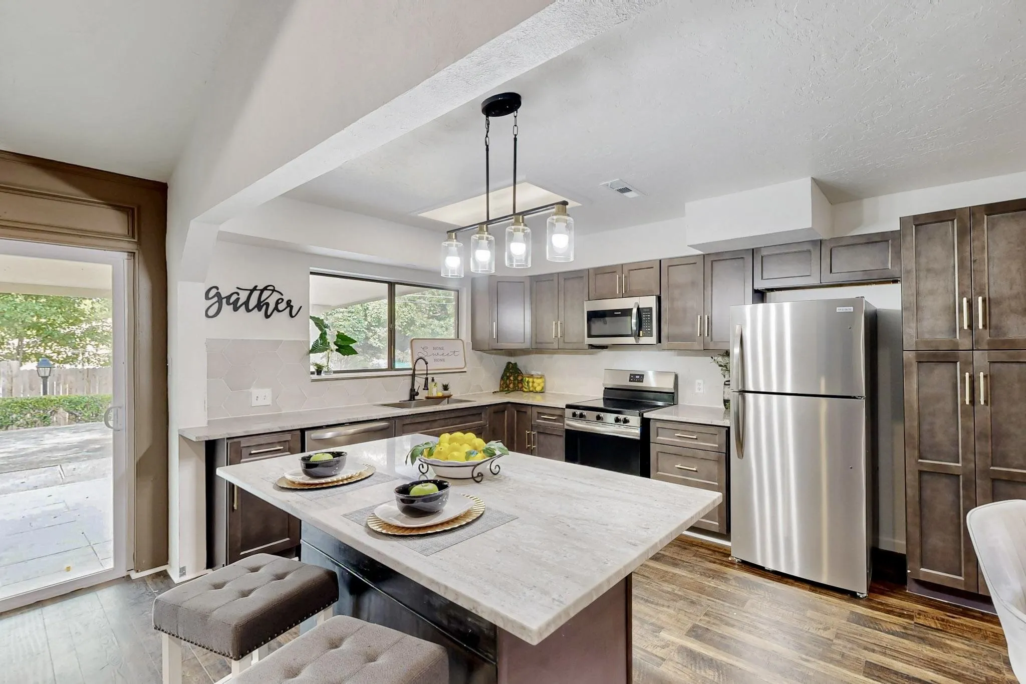 Updated kitchen featuring wood-finish flooring, a central island with a light-toned countertop, stainless steel appliances, dark wood cabinetry, and a sliding glass door to the exterior